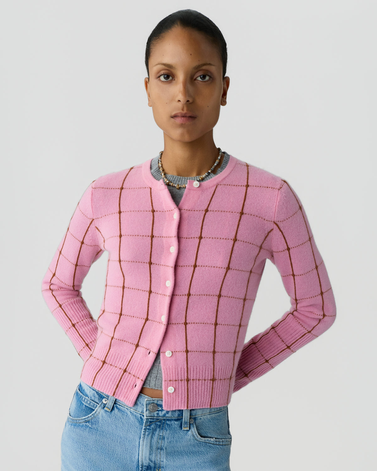 Model wearing a pink checked button front cardigan with long sleeves and light blue jeans while standing against a pale gray background