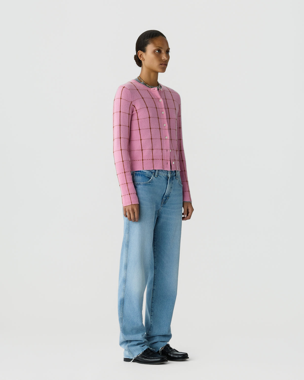 Model wearing a pink checked button cardigan with wide leg blue jeans and black shoes against a plain light background