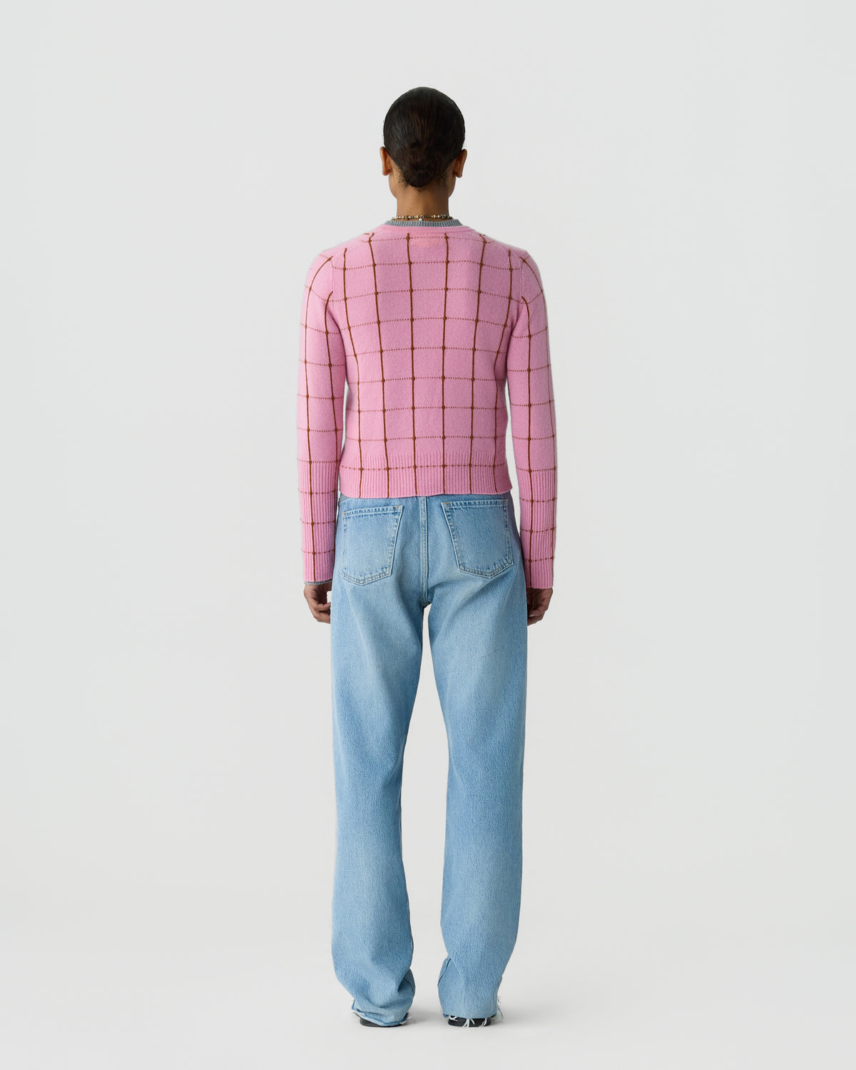 Person seen from behind wearing a pink grid patterned sweater and loose light blue jeans against a plain light gray background