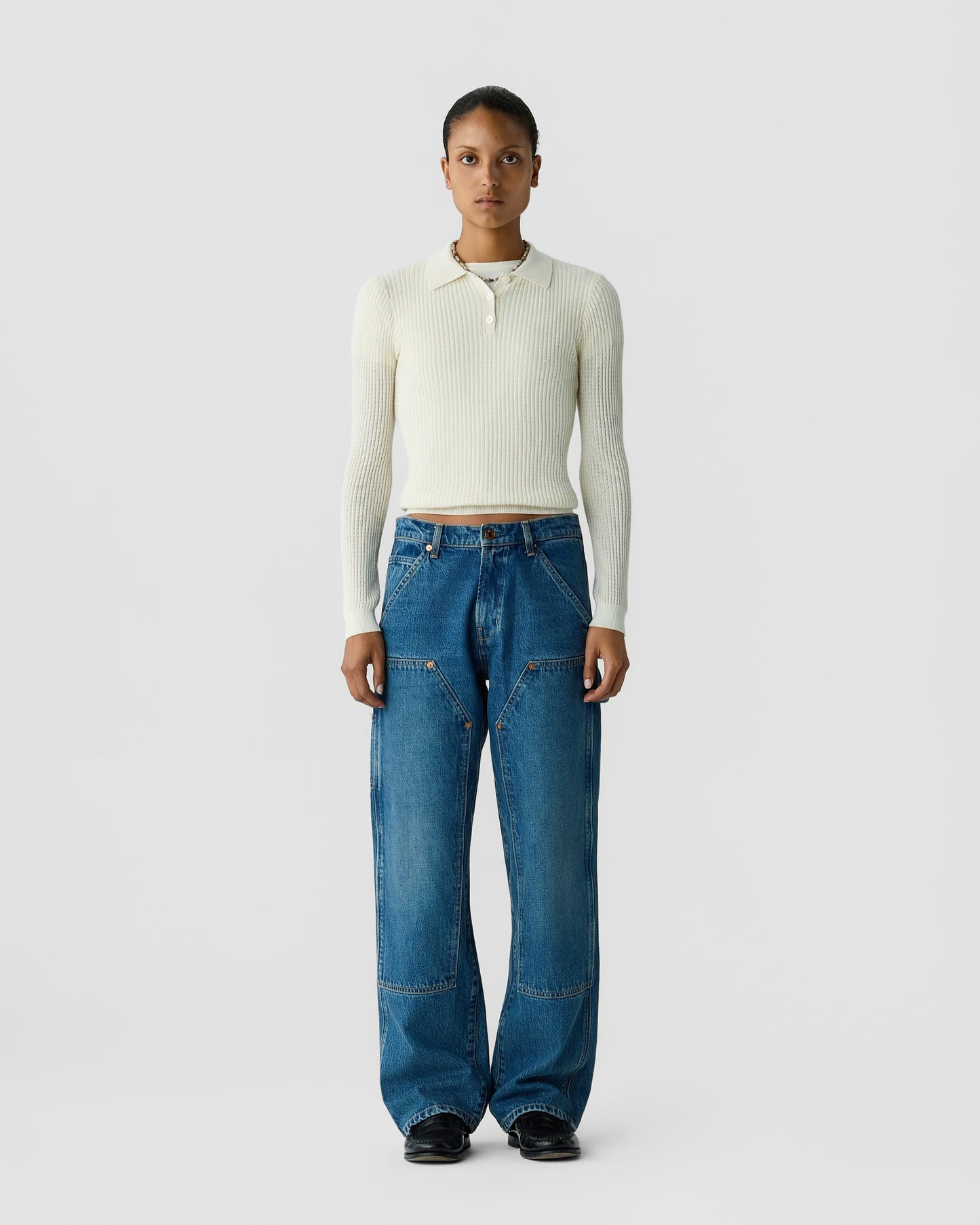 Full-length portrait of a person wearing a cream ribbed sweater, relaxed blue jeans, and black shoes against a light gray background