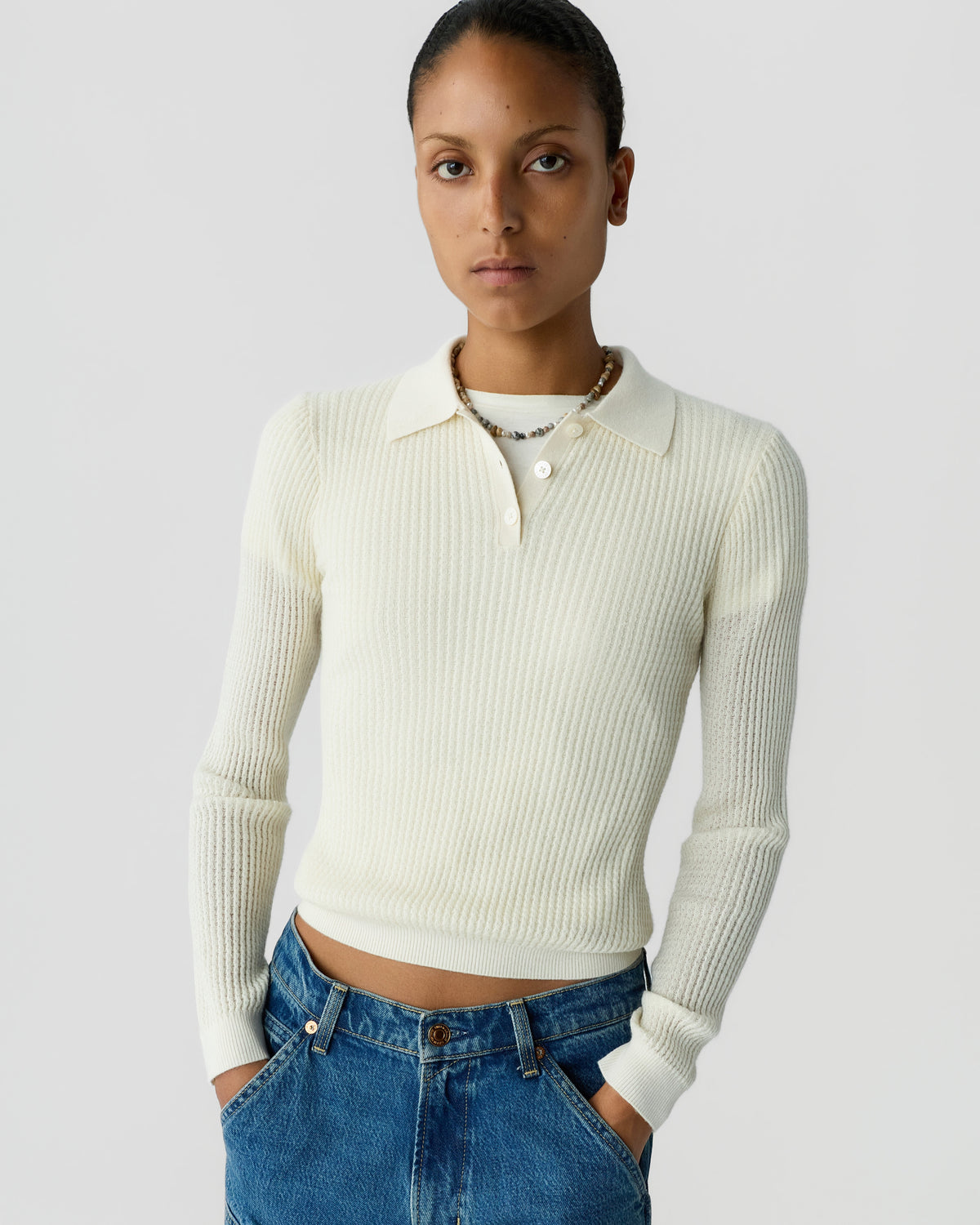 Woman wearing a cream ribbed collared sweater with blue jeans, standing against a light gray background with one hand in a pocket