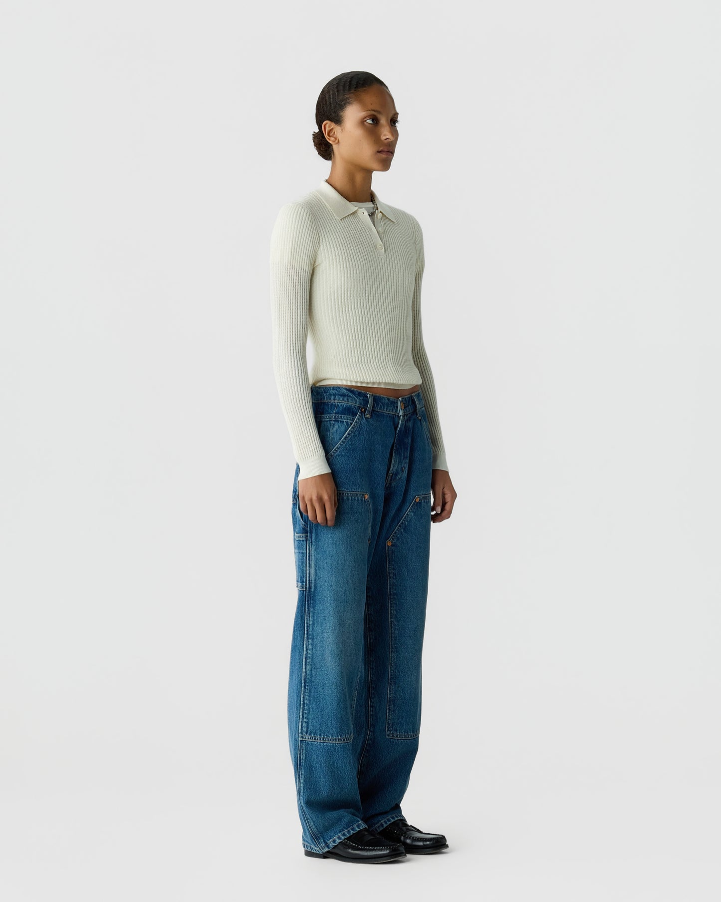 Woman wearing a cream ribbed collared sweater, blue wide-leg jeans, and black shoes against a light gray studio background