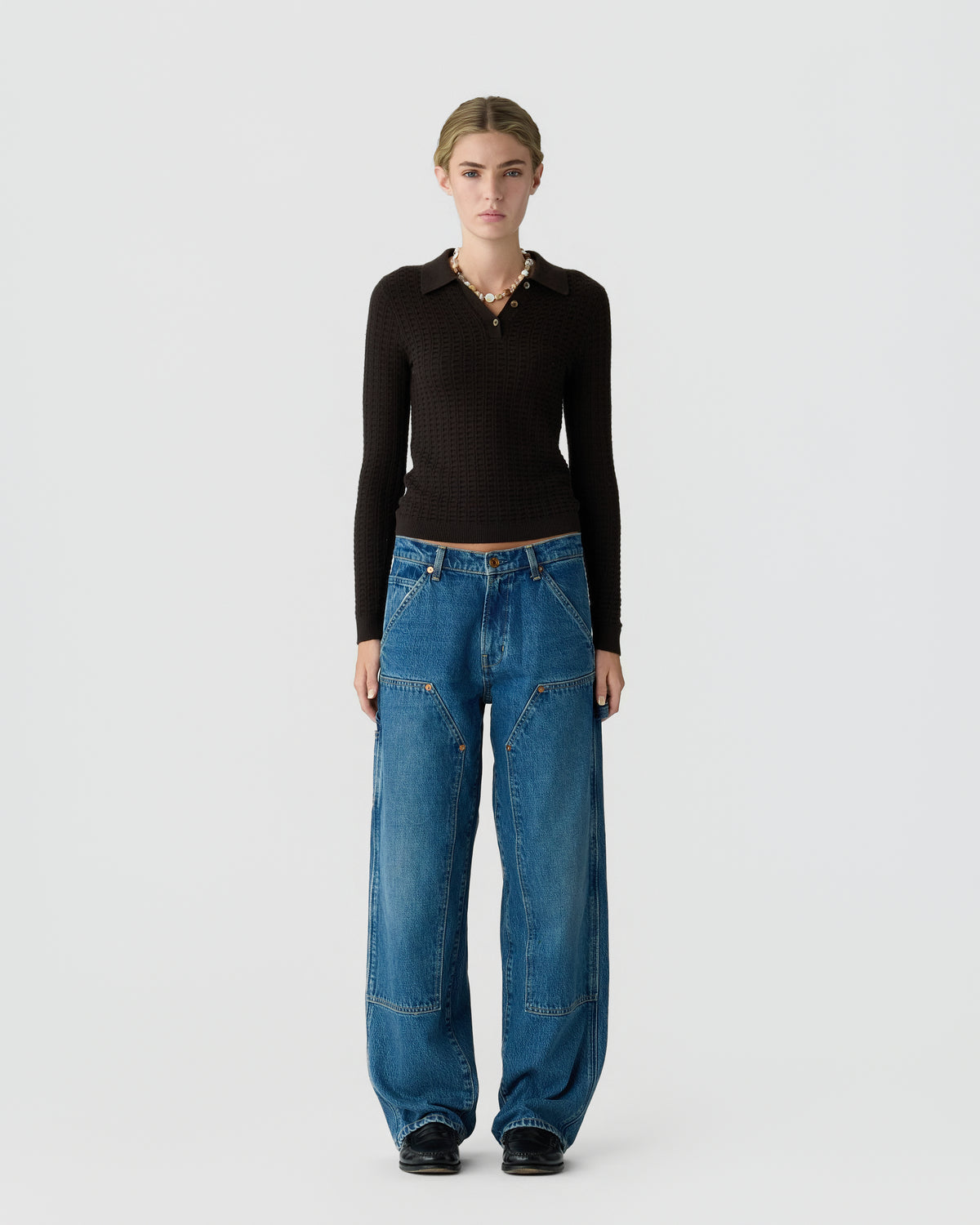 Model standing against a light gray background wearing a black collared knit top, relaxed blue jeans, and black shoes.