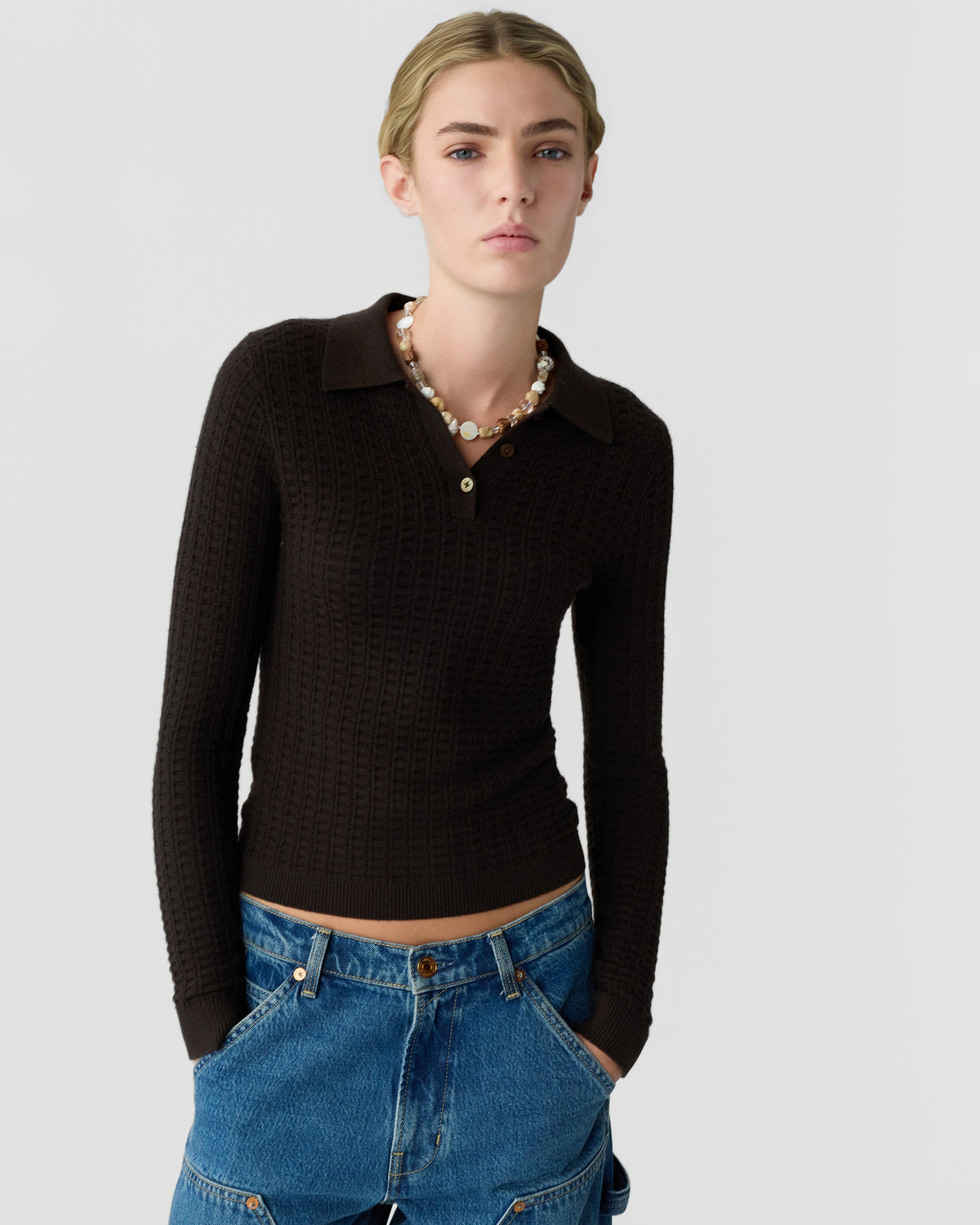 Model wearing a black textured collared knit sweater with long sleeves and blue jeans while standing against a light gray background