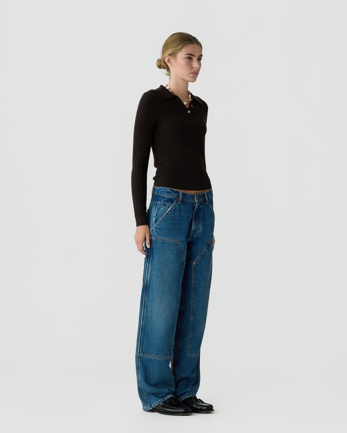 Full-length model wearing a fitted black long-sleeve top, wide-leg blue jeans, and black shoes against a light gray background.