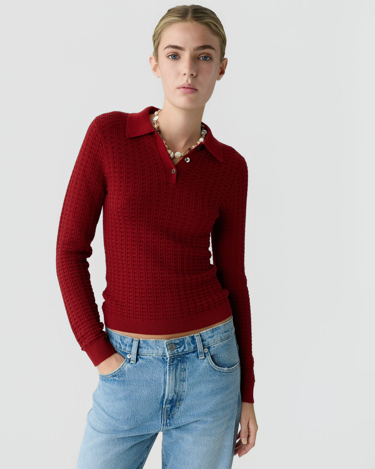 Woman wearing a red collared knit sweater with long sleeves and light blue jeans against a plain light gray background