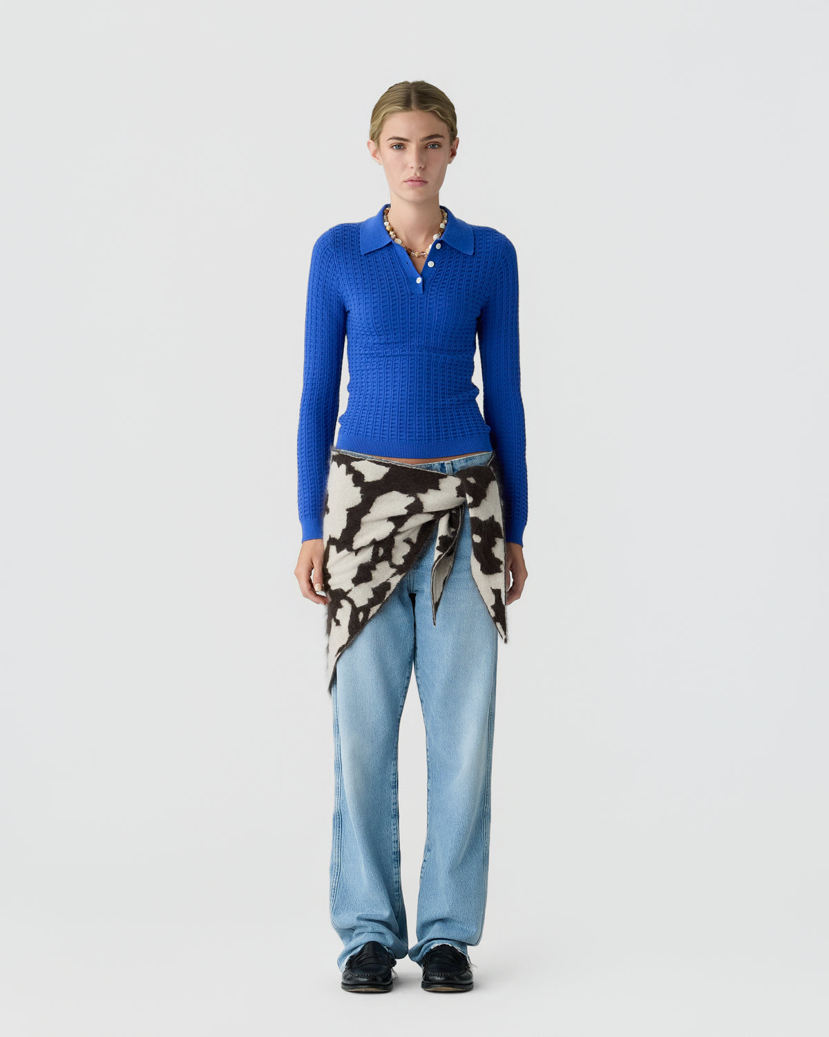 Model wearing a fitted blue collared knit sweater, light blue wide leg jeans, a patterned garment tied at the waist, and black shoes against a pale background