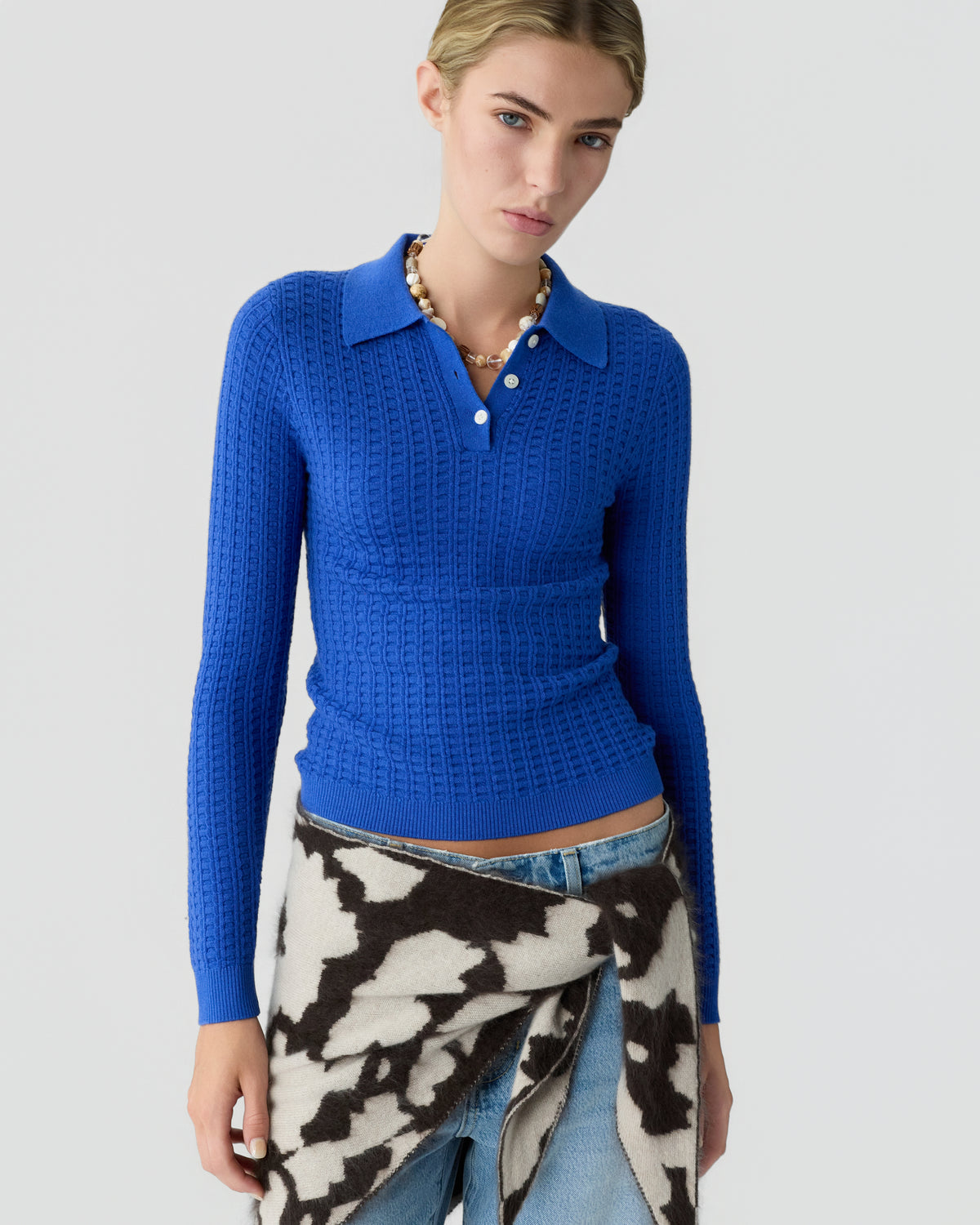 Woman wearing a bright blue textured collared sweater with jeans and a black and white cow print garment tied around her waist against a light gray background
