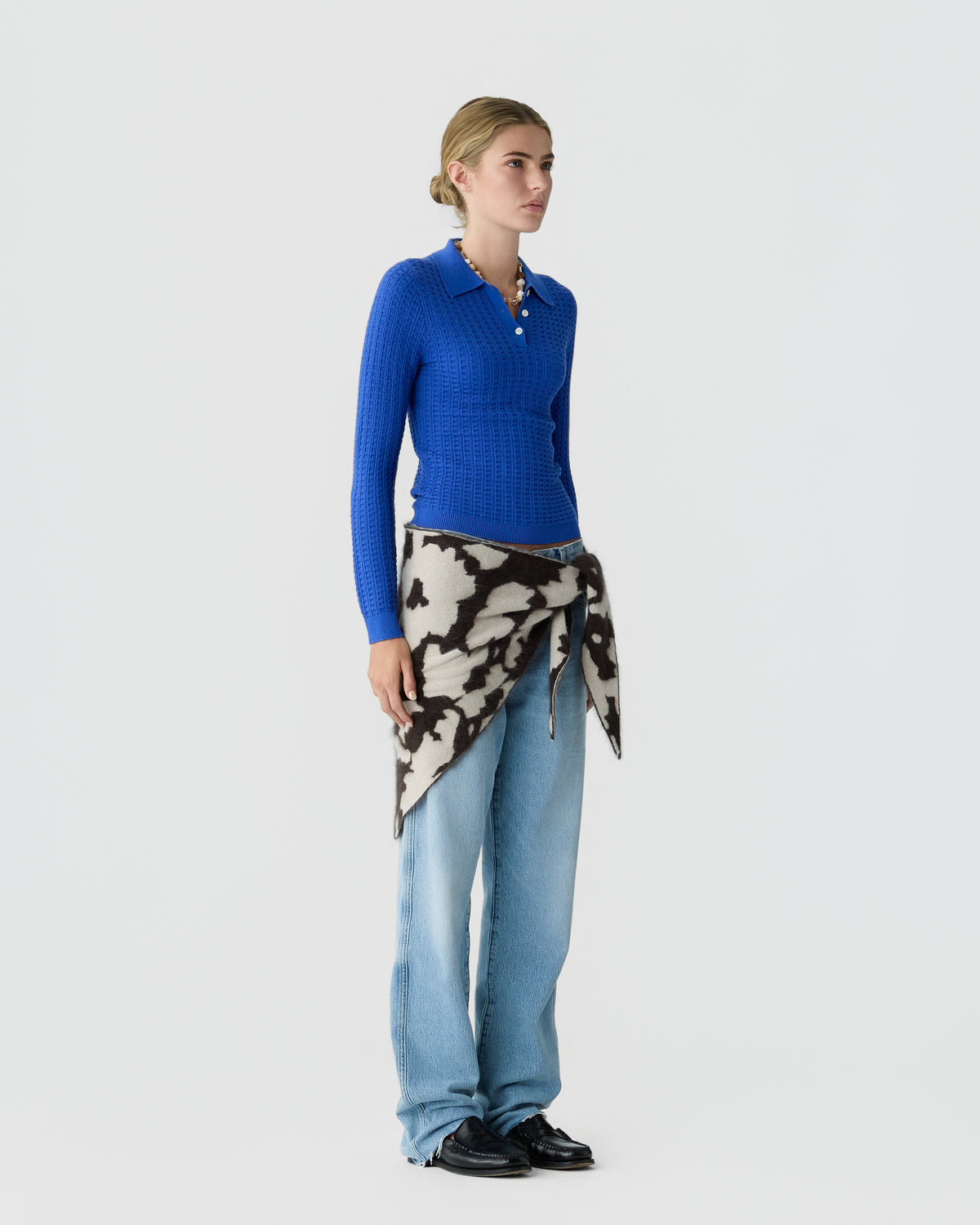 Model wearing a fitted blue knit polo with light wash jeans a black and white patterned sweater tied at the waist and black shoes against a light background