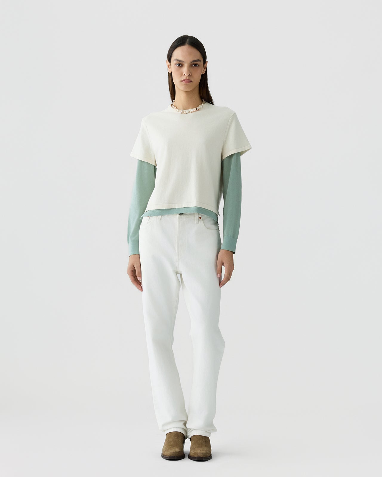 Woman with straight dark hair wearing a layered outfit featuring a cream short-sleeve top over a sage green long-sleeve shirt, paired with white pants and brown shoes against a neutral background.