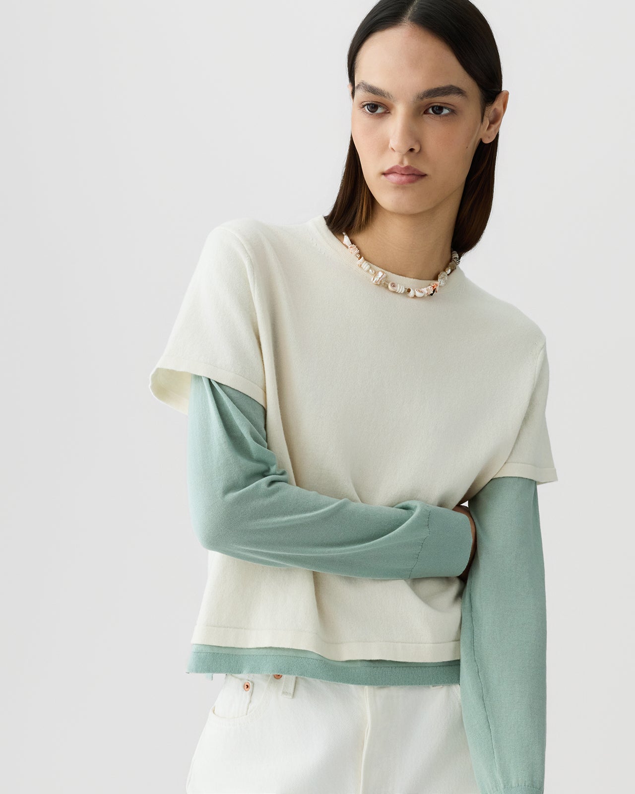 Woman with straight dark hair wearing a cream short-sleeve top layered over a soft sage green long-sleeve shirt, paired with white pants and a shell necklace against a plain light background.
