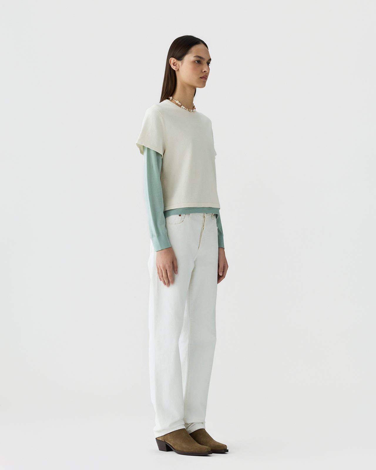 Woman with straight dark hair wearing a cream short-sleeve top layered over a muted sage long-sleeve shirt, paired with relaxed white pants and brown suede ankle boots against a plain light background.