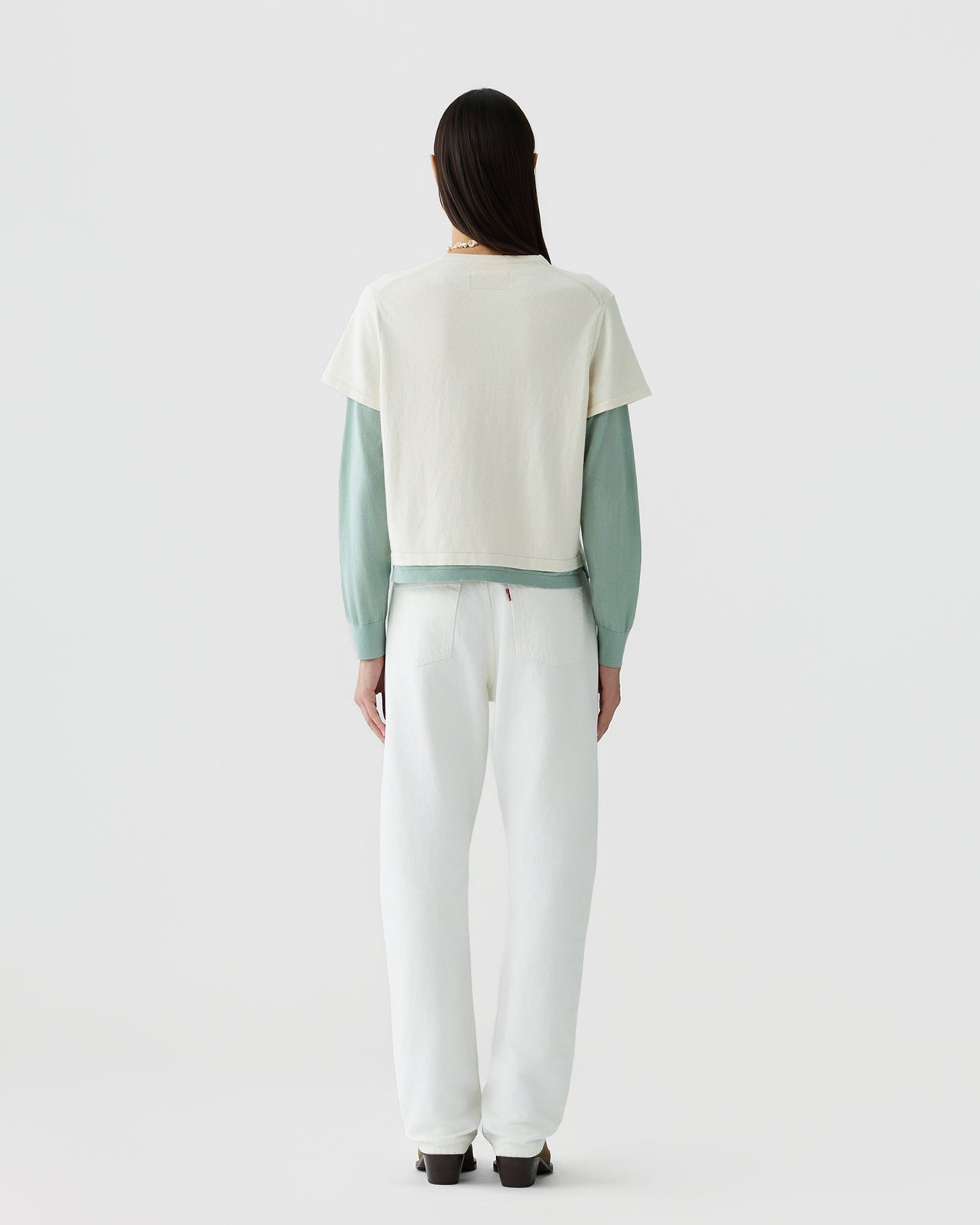 Woman with long dark hair wearing a layered outfit consisting of a cream short-sleeve top over a sage green long-sleeve shirt, paired with loose white pants and dark shoes against a plain light background.