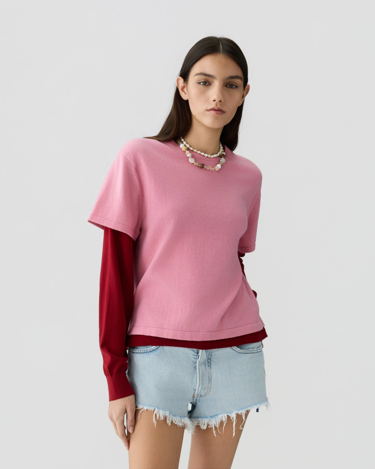 Young woman with long dark hair wearing a soft pink short-sleeve top layered over a deep red long-sleeve shirt, paired with light blue denim shorts with frayed edges, standing against a plain light background.