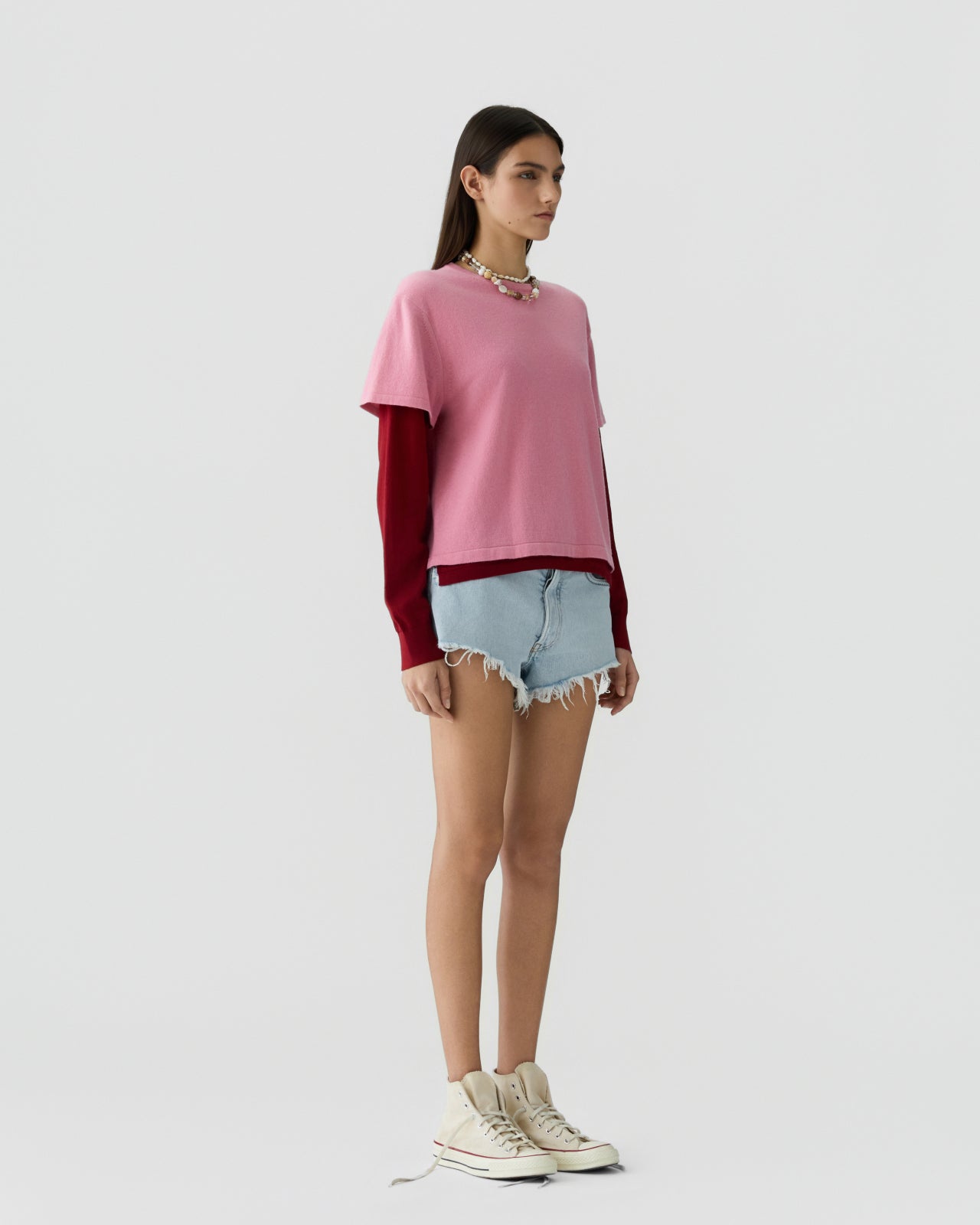 Woman with straight dark hair wearing a layered outfit featuring a soft pink short-sleeve tee over a deep red long-sleeve top, paired with light blue frayed denim shorts and light sneakers against a plain light background.
