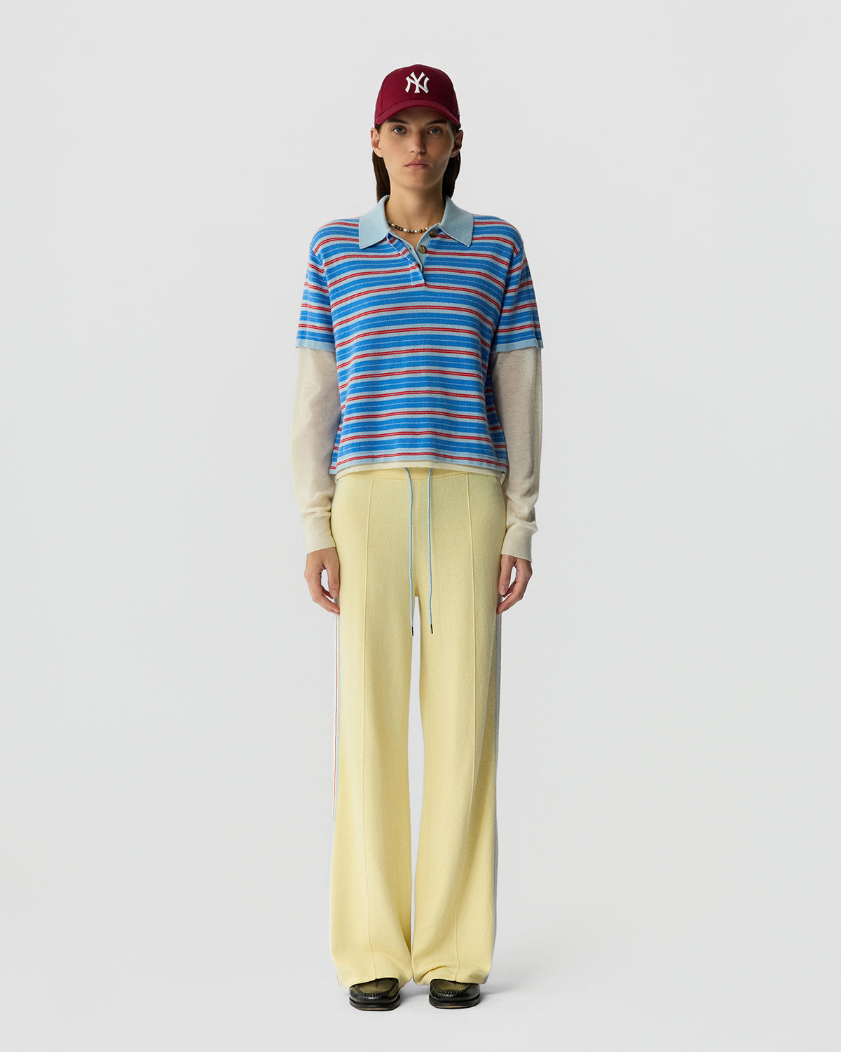 Young model facing forward wearing a blue and red striped short-sleeve polo layered over a cream long-sleeve shirt, paired with pale yellow wide-leg pants and a maroon baseball cap.