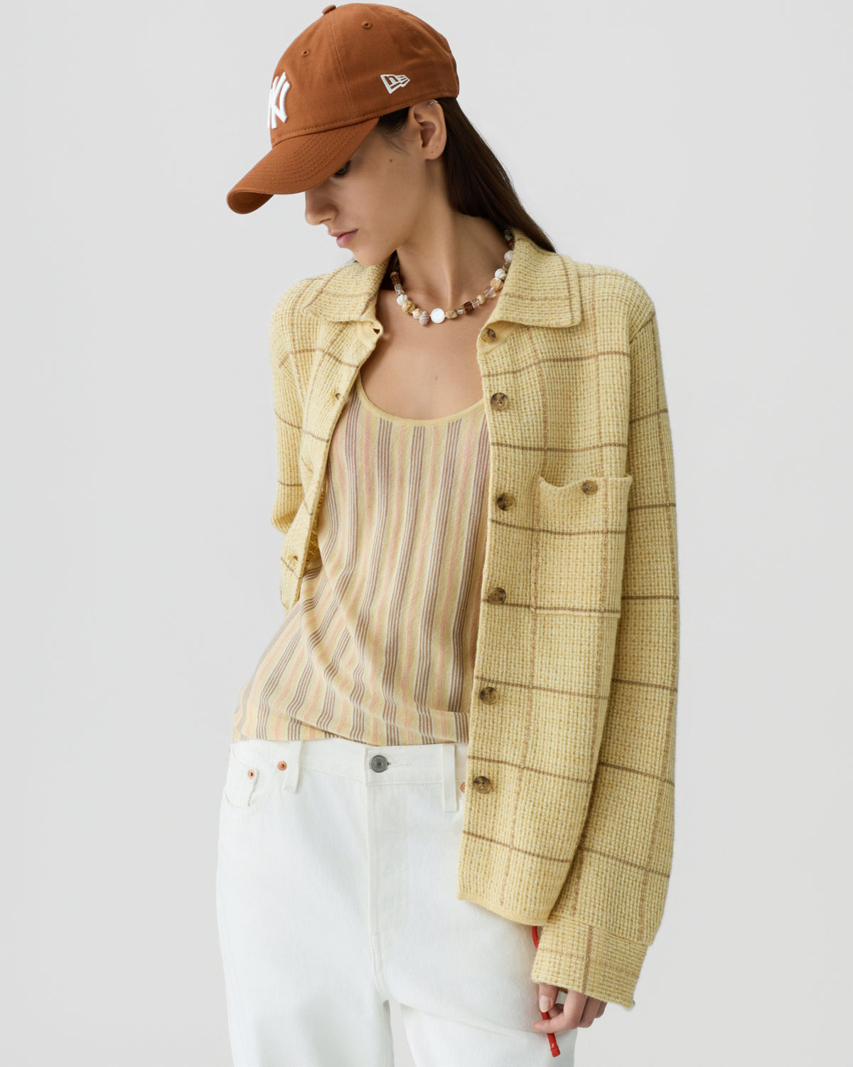 Woman wearing a pale yellow checkered knit jacket over a beige striped tank top, paired with high-waisted white jeans and a rust-colored baseball cap, posing against a light background.