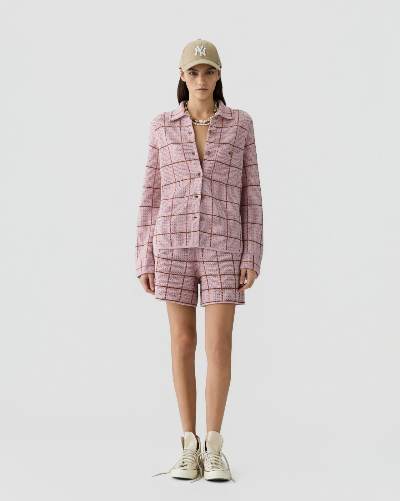 Woman wearing a pink knit shirt and matching shorts featuring a brown grid pattern, paired with a beige baseball cap, chunky sneakers, and a layered necklace against a plain background.