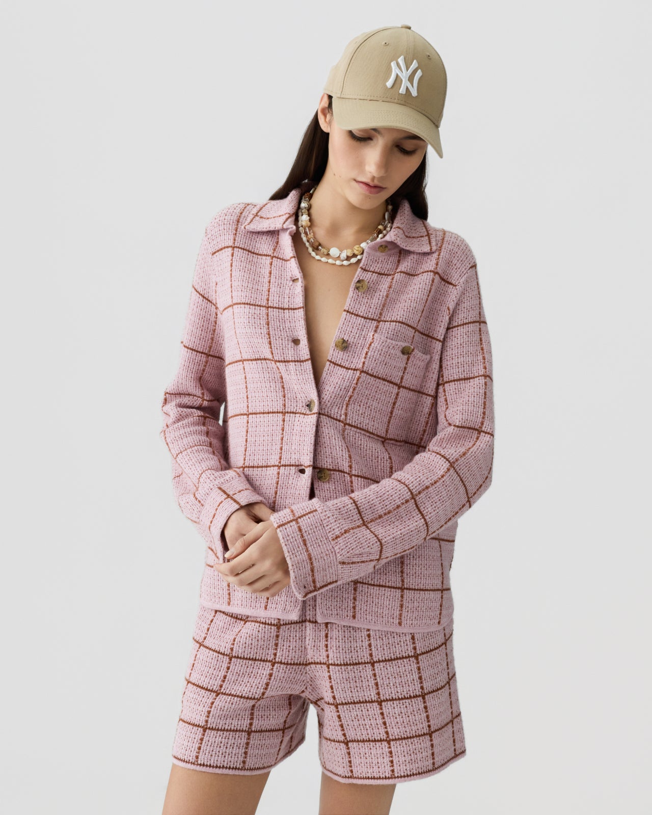 Woman wearing a textured soft pink knit jacket and matching shorts with a brown windowpane pattern, accessorized with a beige baseball cap and a layered pearl necklace.