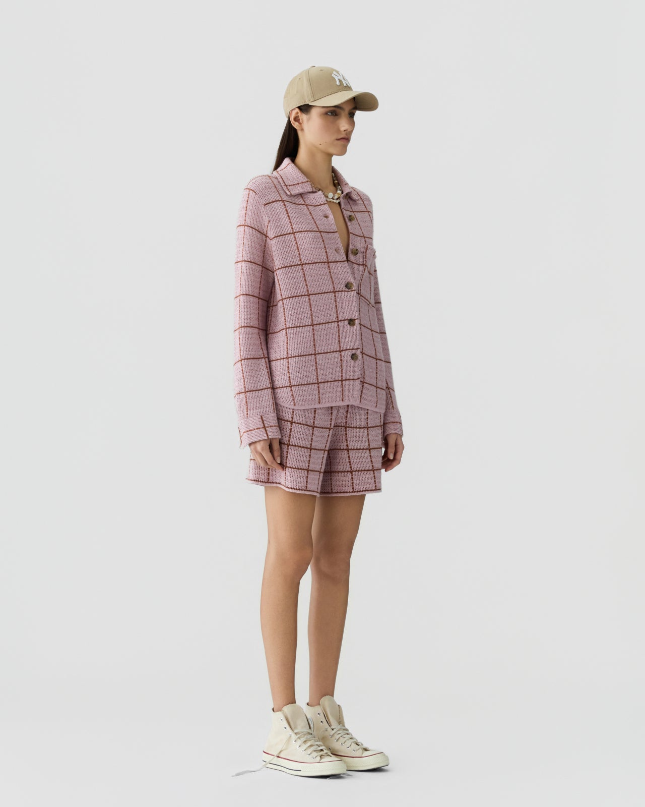Woman standing in profile wearing a soft pink knit jacket and matching shorts with a brown grid pattern, paired with cream high-top sneakers and a beige baseball cap.