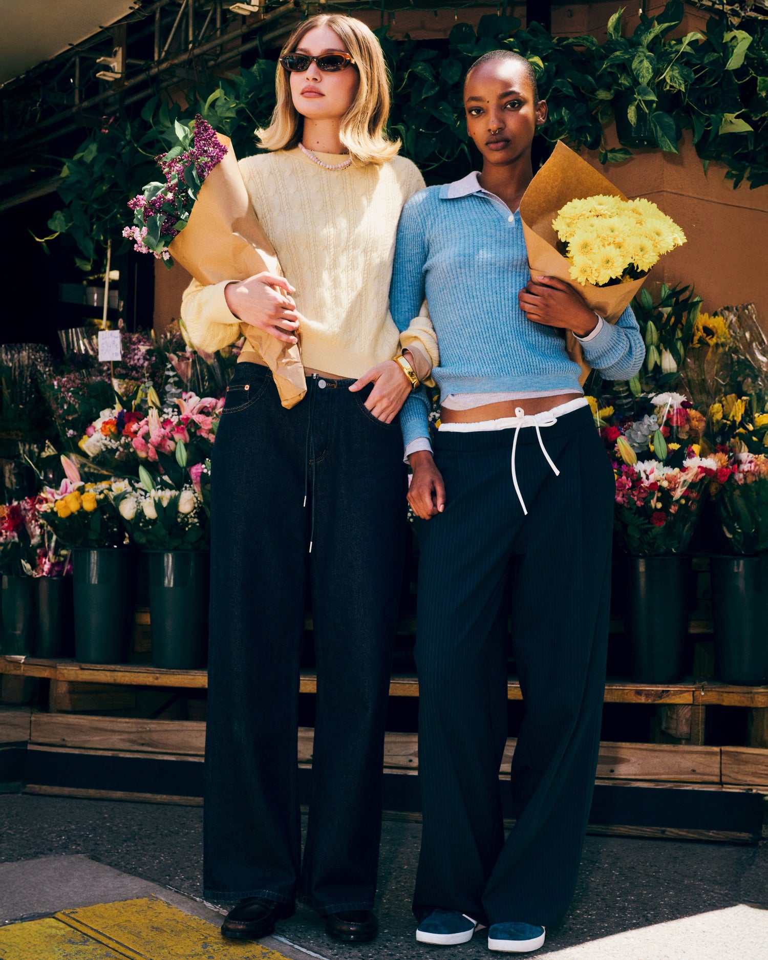 Two women standing outdoors in front of a flower shop, each holding a bouquet; one wearing a cream knit sweater and sunglasses, the other in a light blue sweater with dark wide-leg pants.
