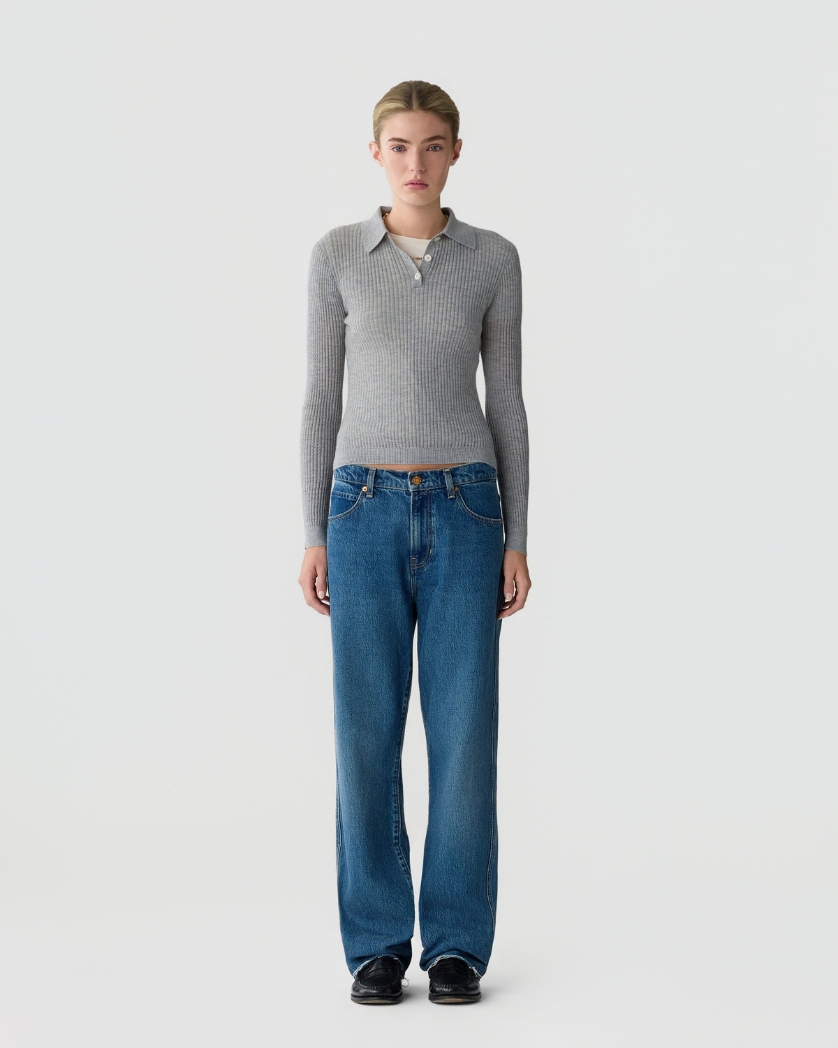 Model wearing a fitted gray collared knit sweater, blue wide-leg jeans, and black shoes while standing against a plain light gray studio background