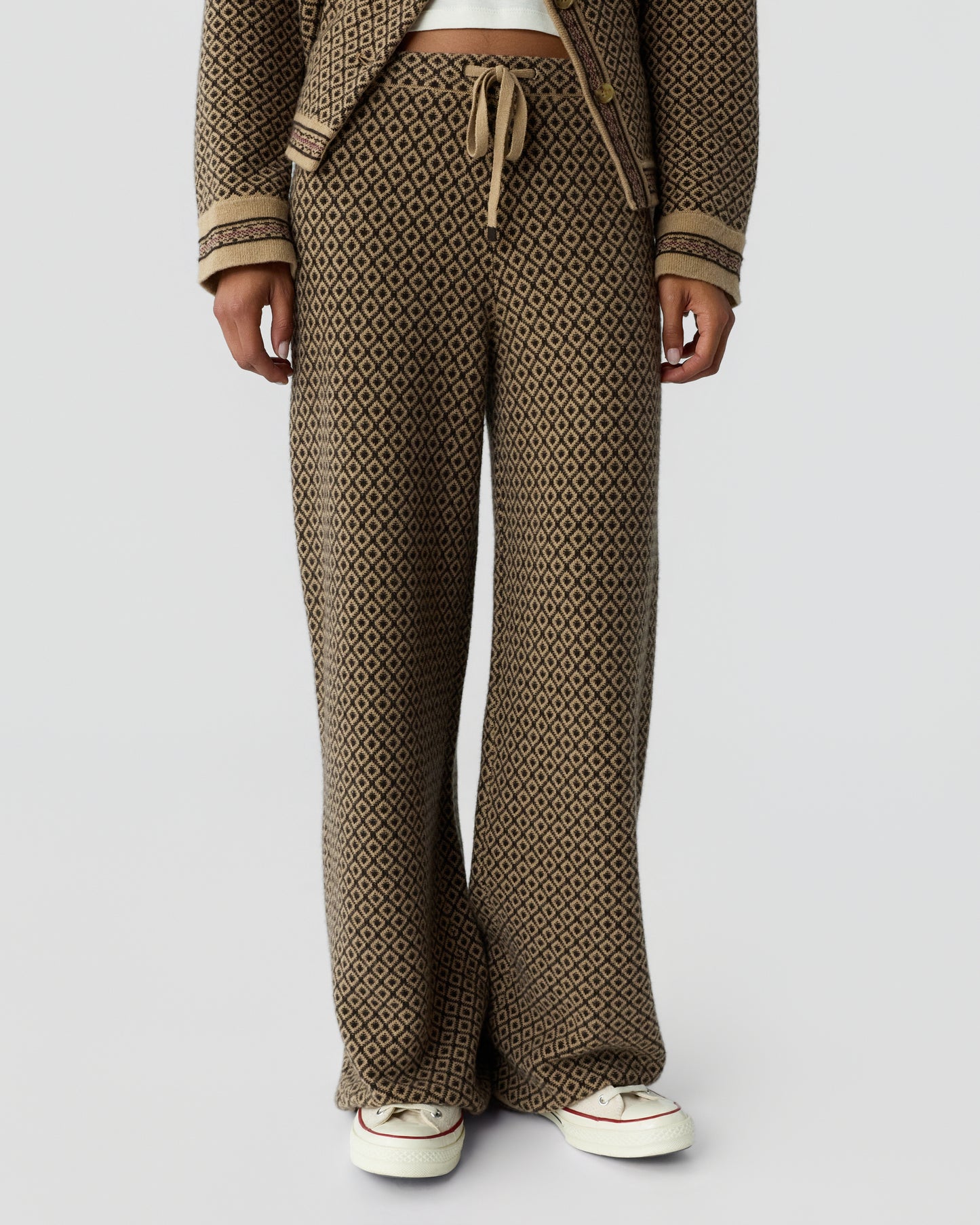 Close-up of a person wearing wide-leg pants with a brown and black diamond pattern and a drawstring waist, paired with white sneakers and a matching cardigan.