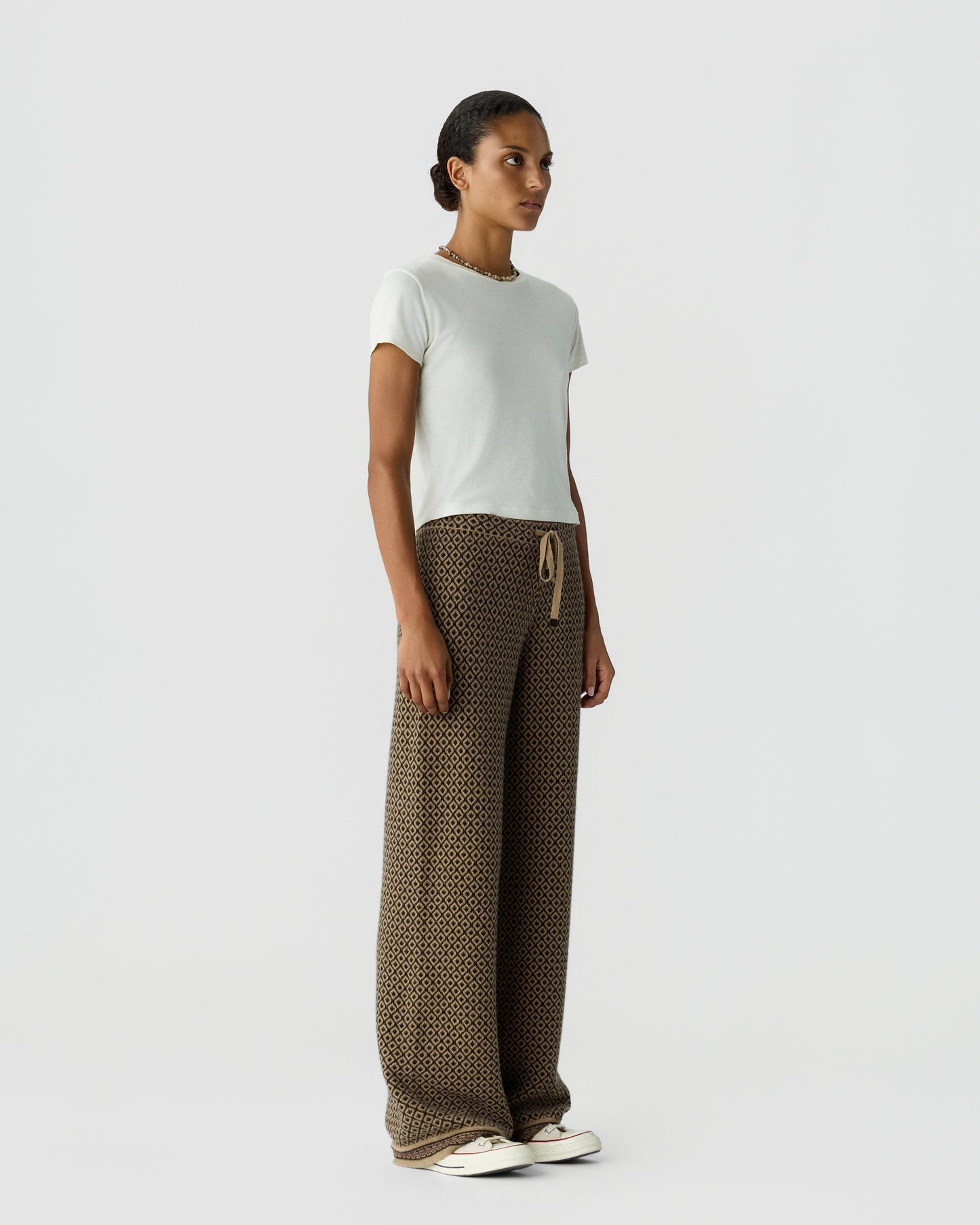 Woman standing against a plain background wearing a white short-sleeve top, patterned wide-leg pants with a drawstring waist, and white casual shoes.