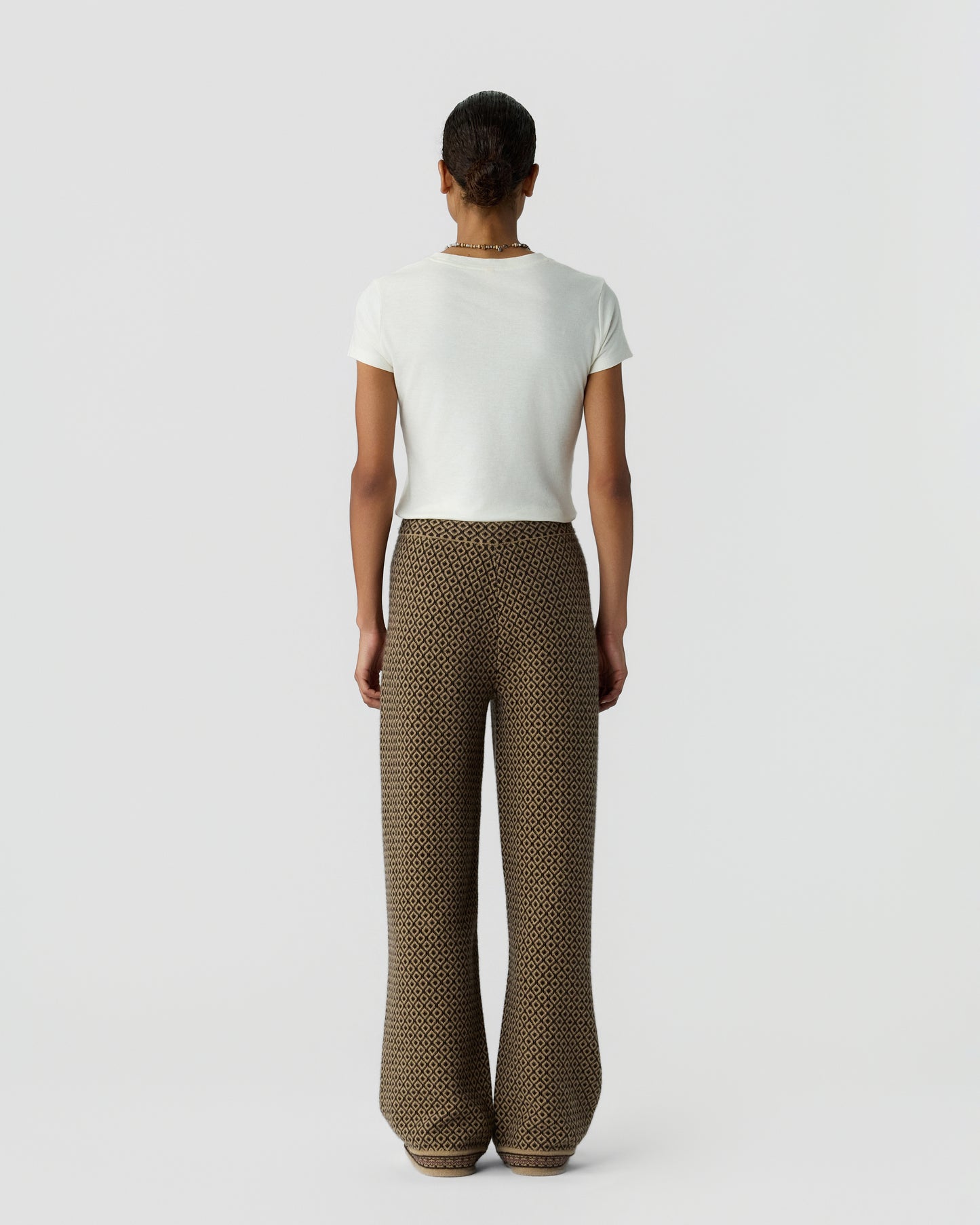 Person standing facing away wearing a plain white short-sleeve top and high-waisted wide-leg pants with a brown and black geometric pattern against a white background.