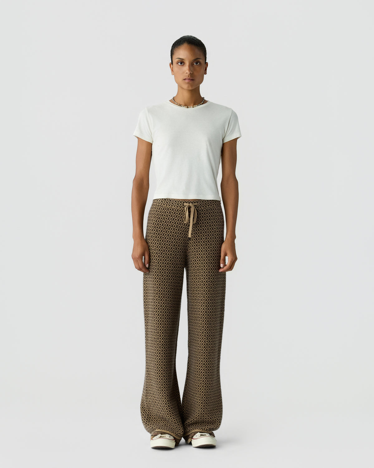 Woman standing against plain background wearing a white short-sleeve top and wide-legged patterned brown pants with a drawstring waist and white sneakers.