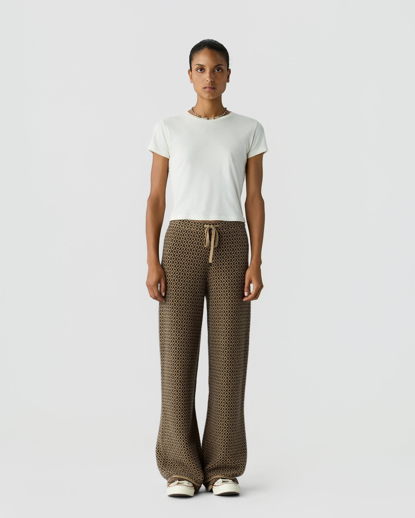 Woman standing against plain background wearing a white short-sleeve top and wide-legged patterned brown pants with a drawstring waist and white sneakers.