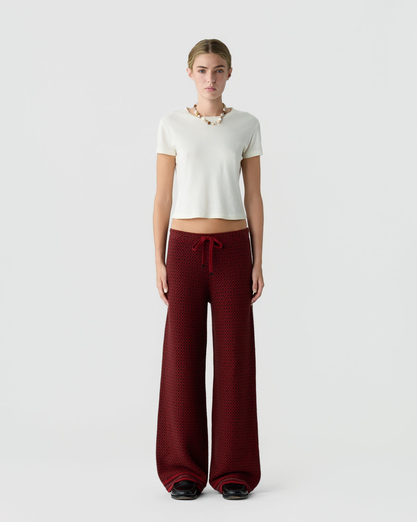 Young woman standing against a plain background wearing a white short-sleeve top and wide-leg red patterned pants with a drawstring waist and black shoes.