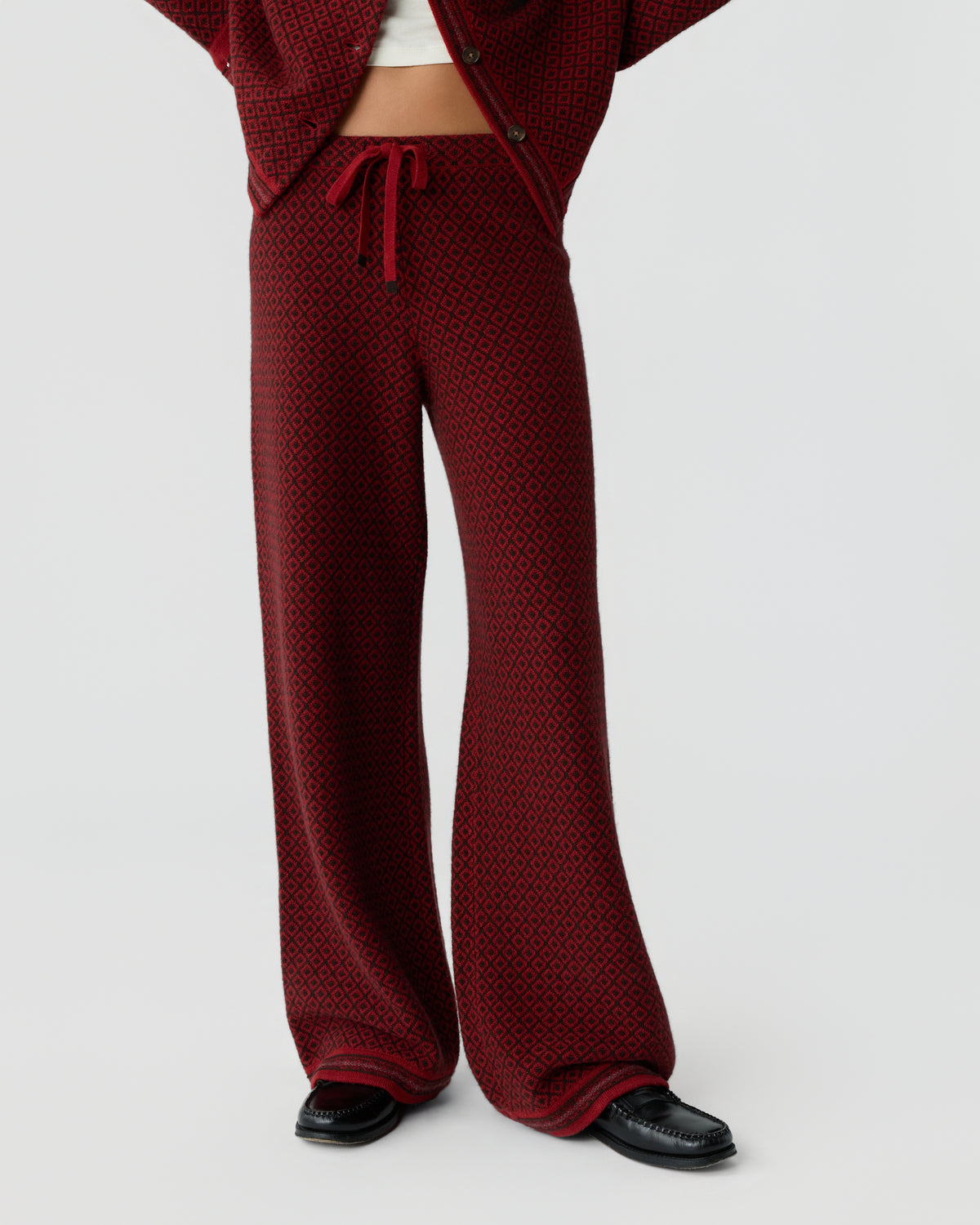 Lower body view of a person wearing wide-legged, red patterned pants with a drawstring waist and black shoes, paired with a matching jacket partially open to show a white top underneath.