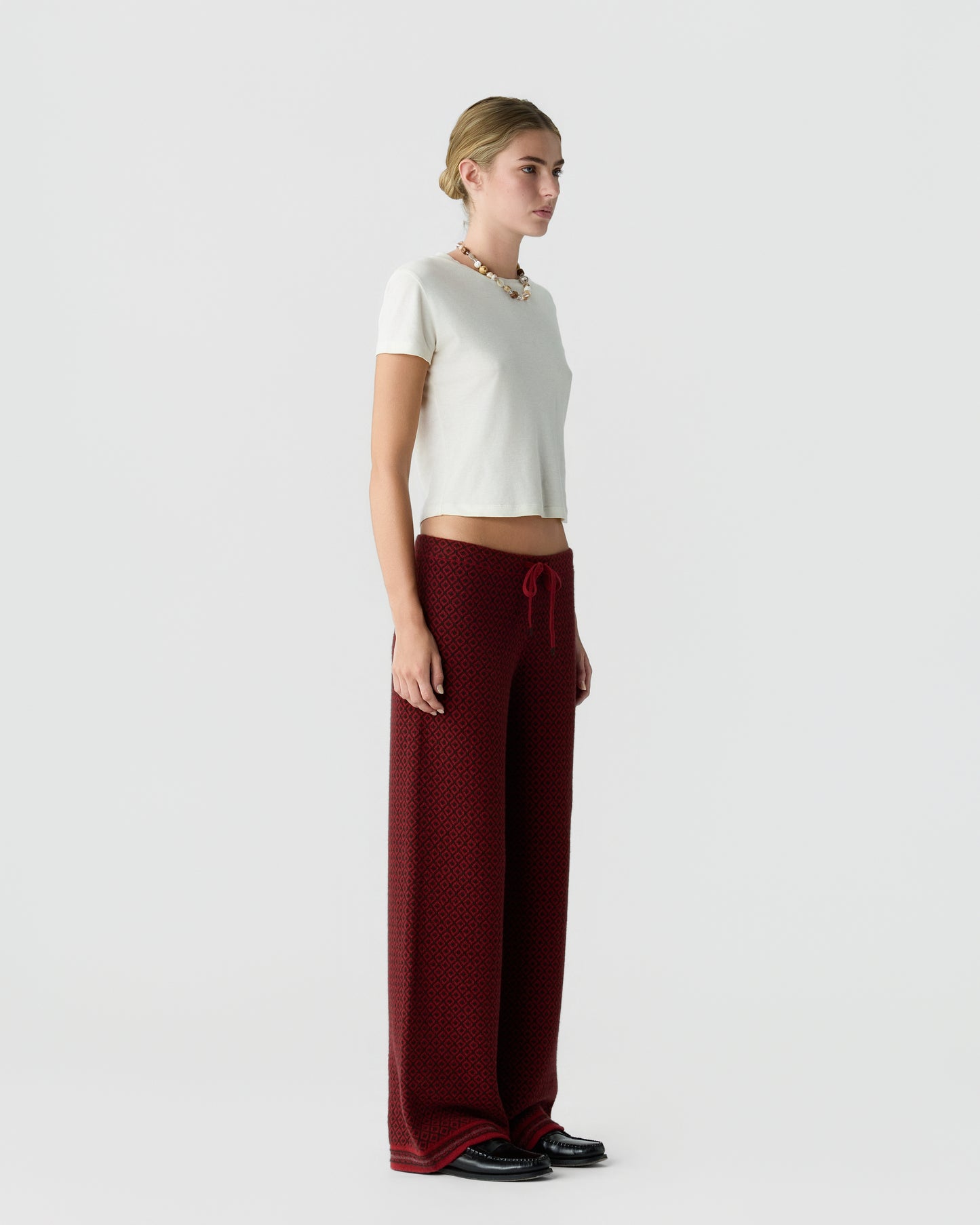 Woman wearing a white short-sleeve top and high-waisted, wide-leg burgundy pants with a textured pattern, standing in profile against a plain white background.