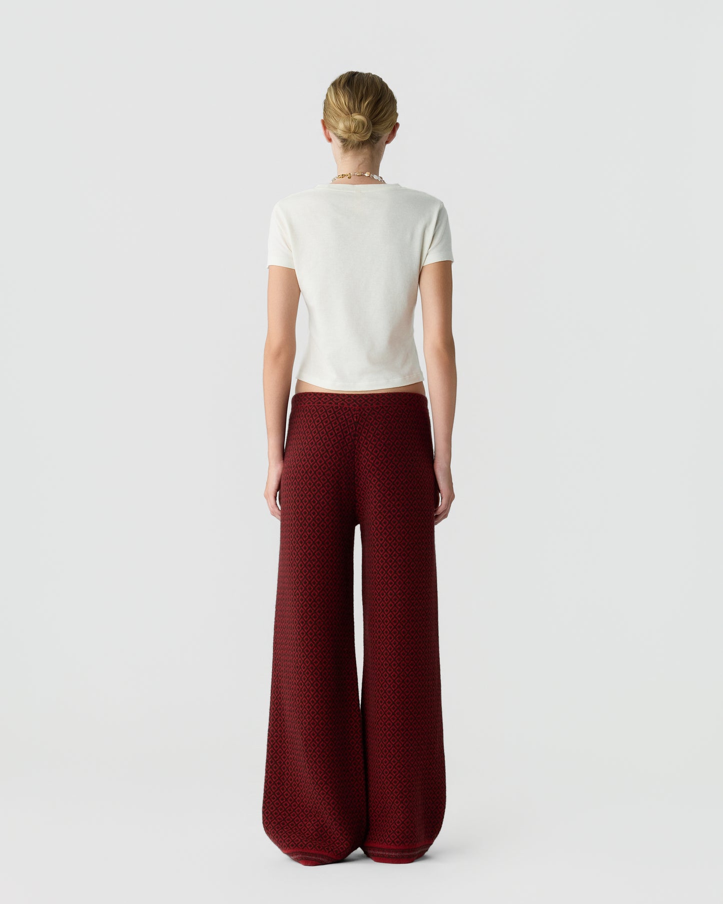 Woman standing facing away wearing a short-sleeve white top and wide-leg maroon pants with a small black pattern against a plain light gray background.