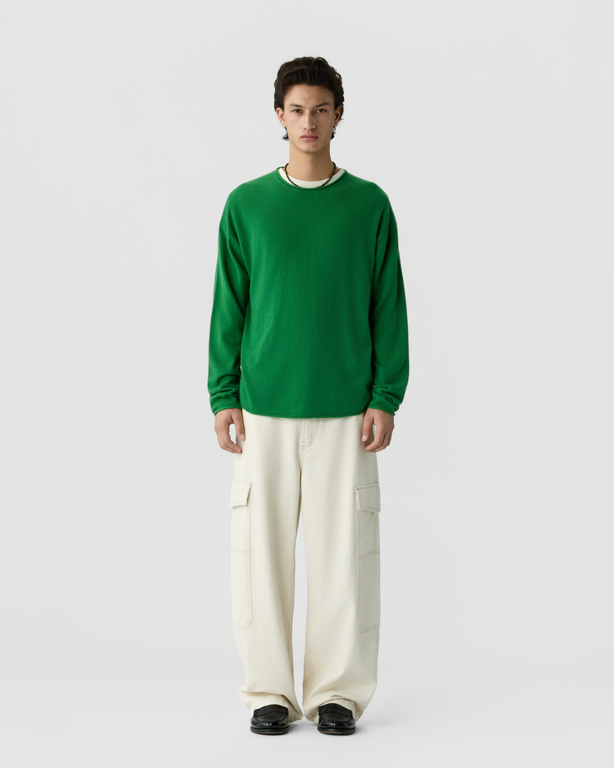 A young man with dark hair stands against a plain light gray background wearing a bright green long-sleeve sweater and loose-fitting off-white cargo pants with black shoes.