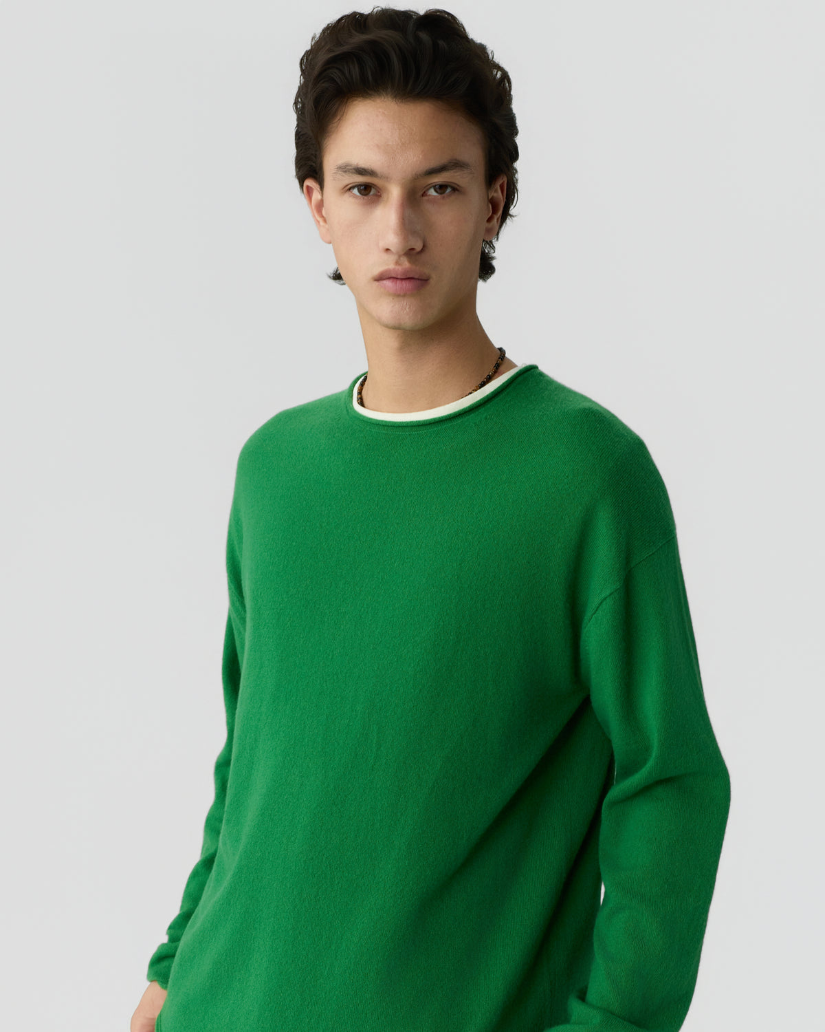 Young man with dark hair wearing a bright green long-sleeve cashmere sweater over a white crew-neck shirt against a plain light background.