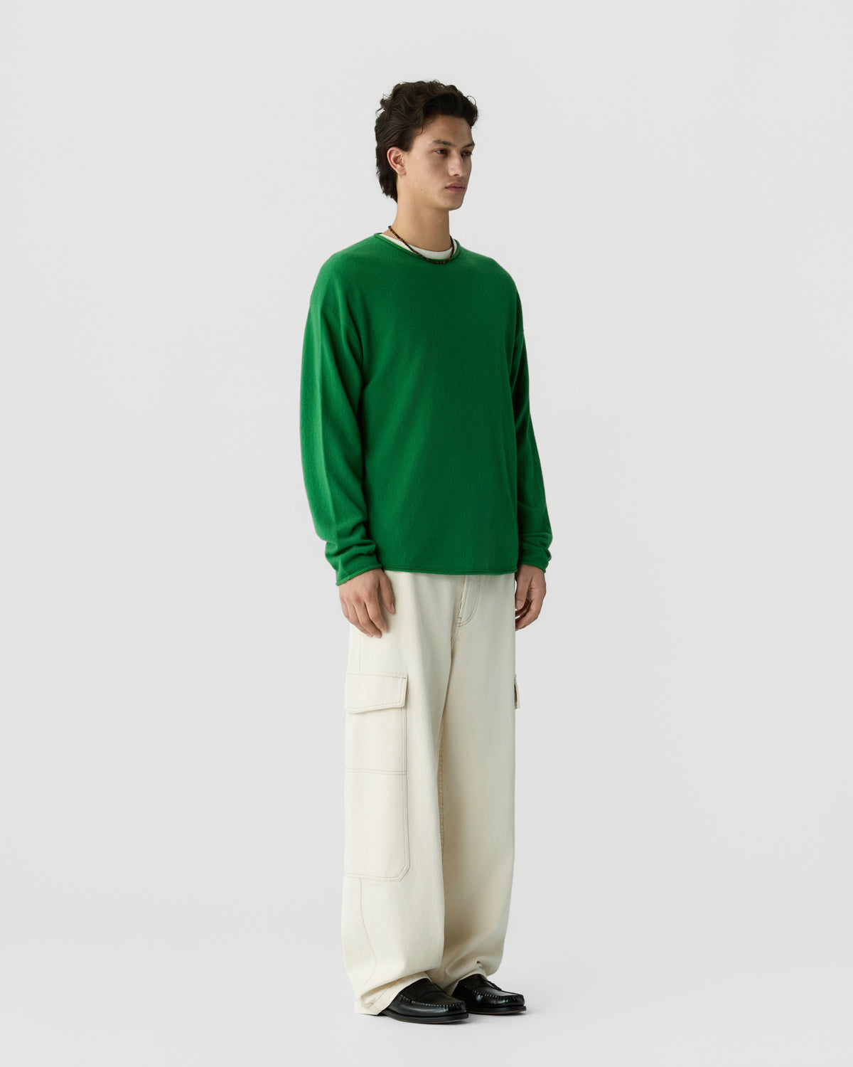 Man standing against a plain background wearing a green long-sleeve cashmere shirt, loose cream-colored cargo pants, and black shoes, looking forward.