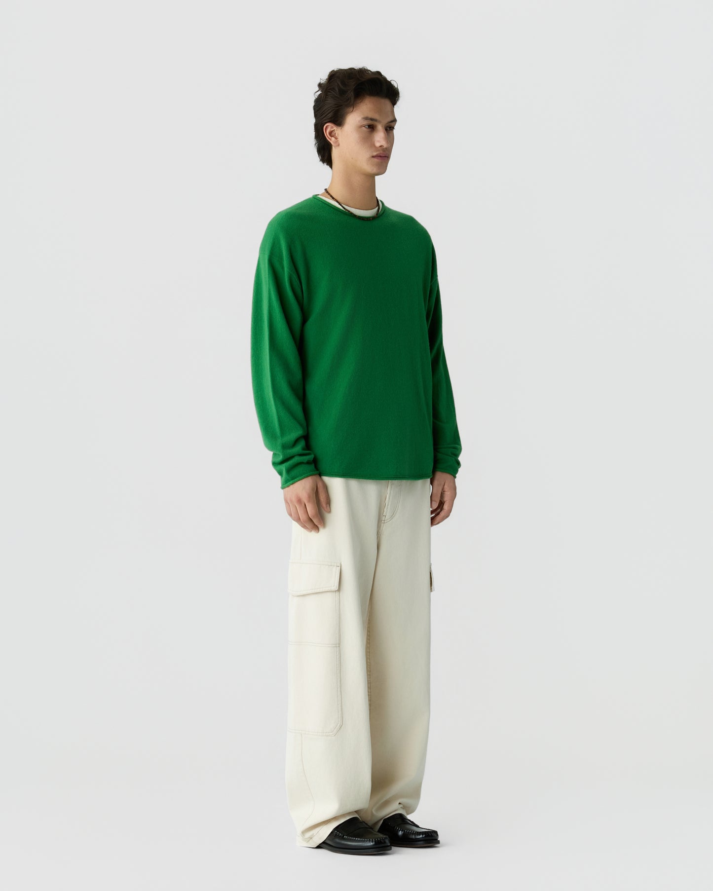 Man standing against a plain background wearing a green long-sleeve cashmere shirt, loose cream-colored cargo pants, and black shoes, looking forward.