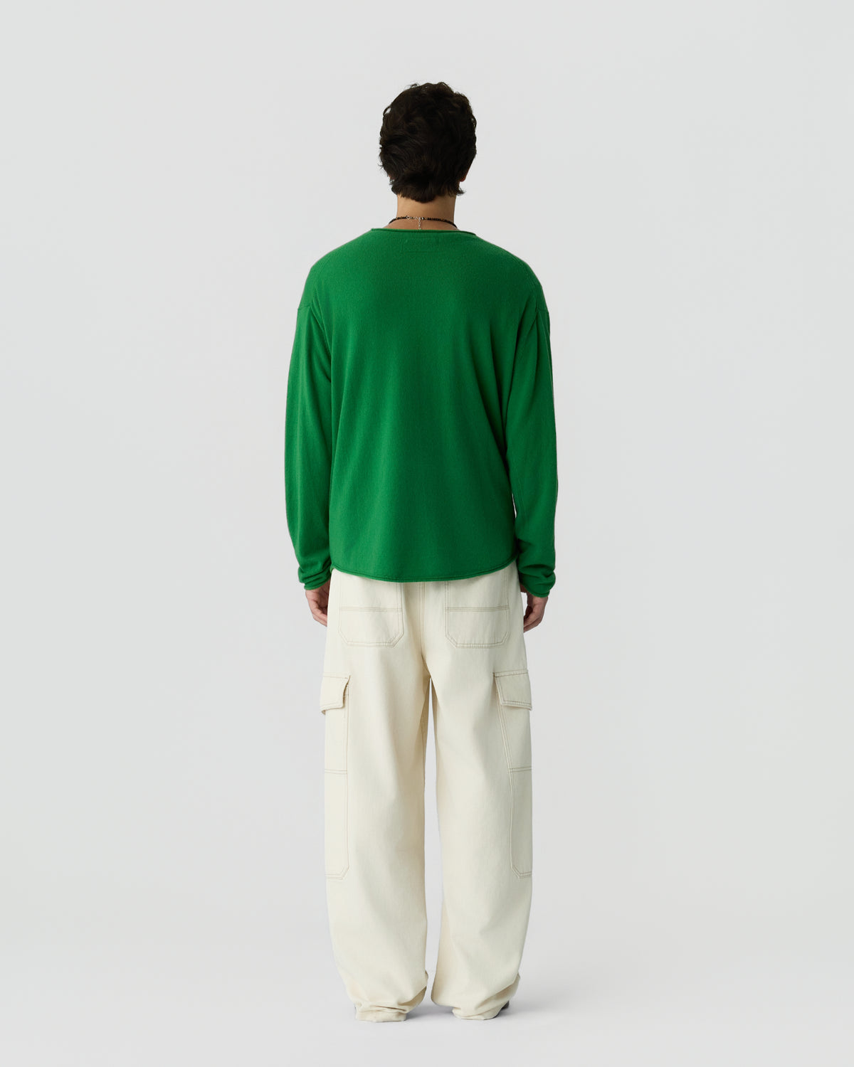 Man wearing a bright green long-sleeve top and loose-fitting off-white cargo pants standing with his back to the camera against a plain light gray background.