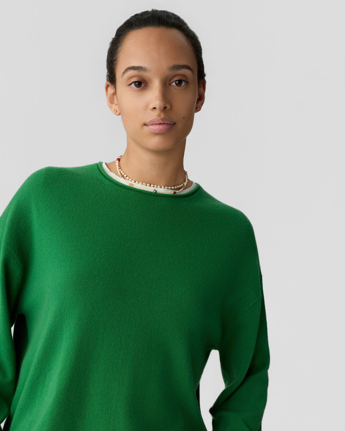 Young woman with dark hair pulled back wearing a green cashmere sweater and a beaded necklace against a plain light gray background