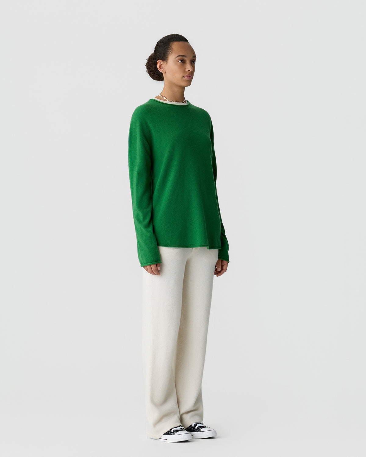 Woman standing against a plain light background wearing a green long-sleeve sweater, white wide-leg pants, and black and white sneakers, looking to the side.
