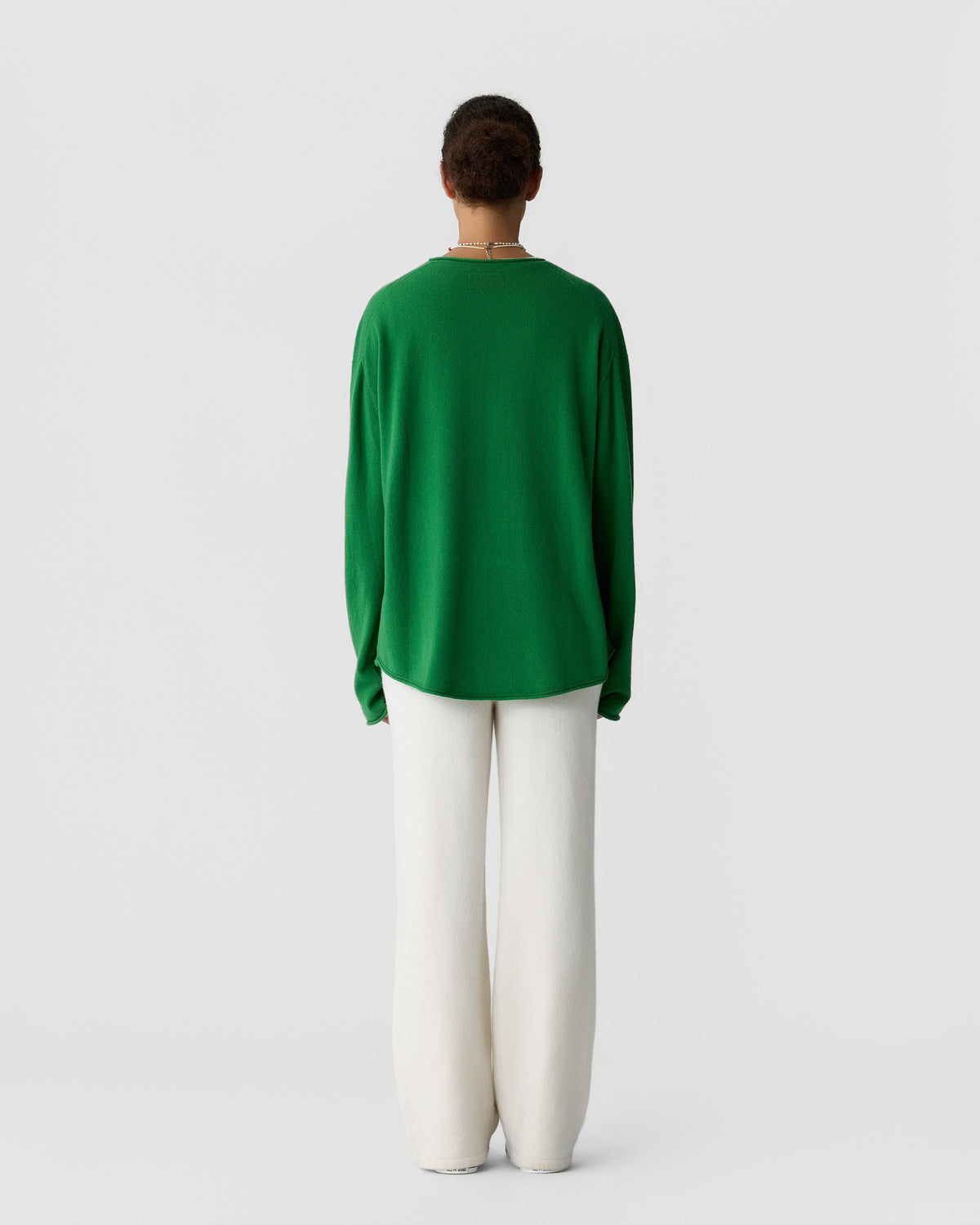 Person wearing a long-sleeve green top and loose white pants standing with their back to the camera against a plain light background