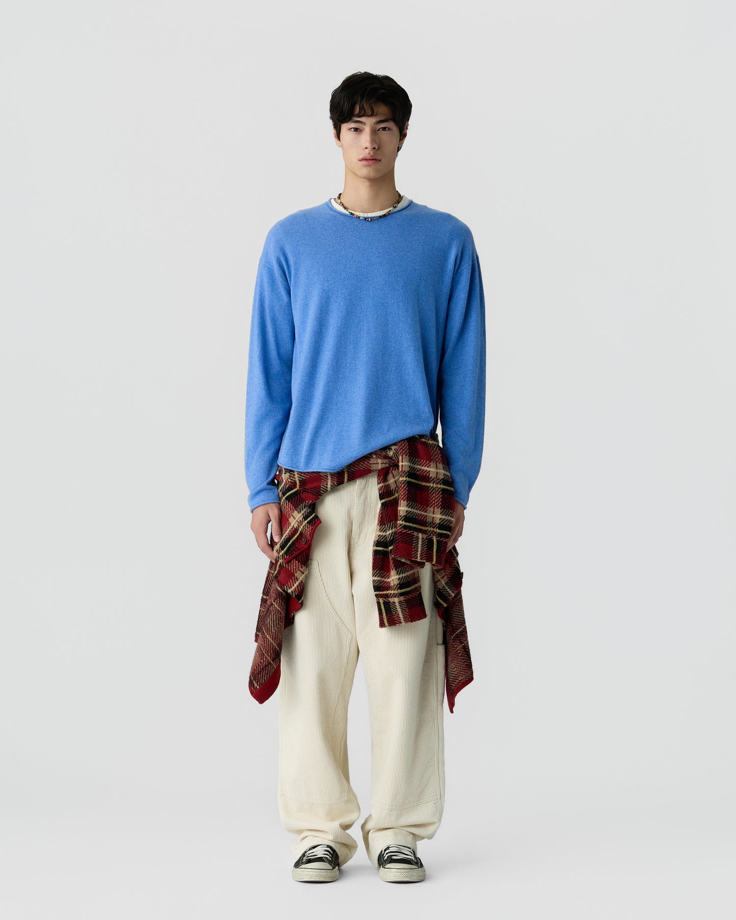 Young man wearing a bright blue sweater, cream wide-leg pants, black and white sneakers, and a red plaid shirt tied around his waist, standing against a plain white background.