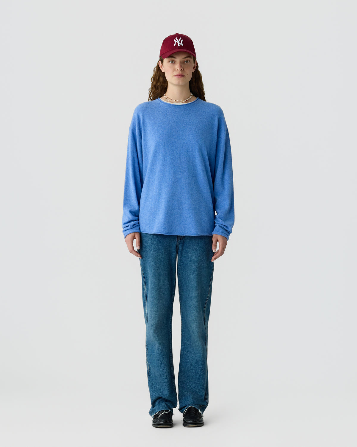 Woman wearing a loose-fitting blue long-sleeve cashmere sweater, straight-leg blue jeans, black shoes, and a red baseball cap standing against a plain light background.