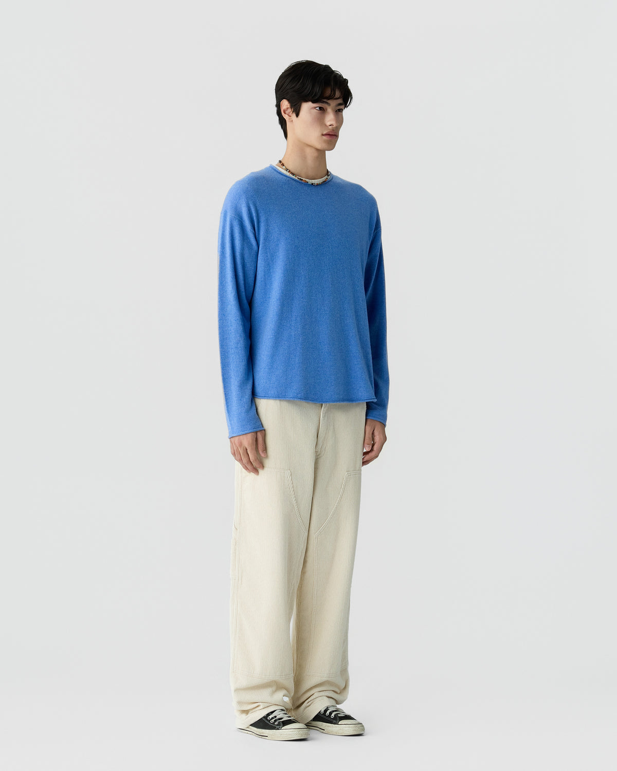 Young man wearing a blue long-sleeve sweater with loose cream-colored pants and black and white sneakers, standing against a plain light background.