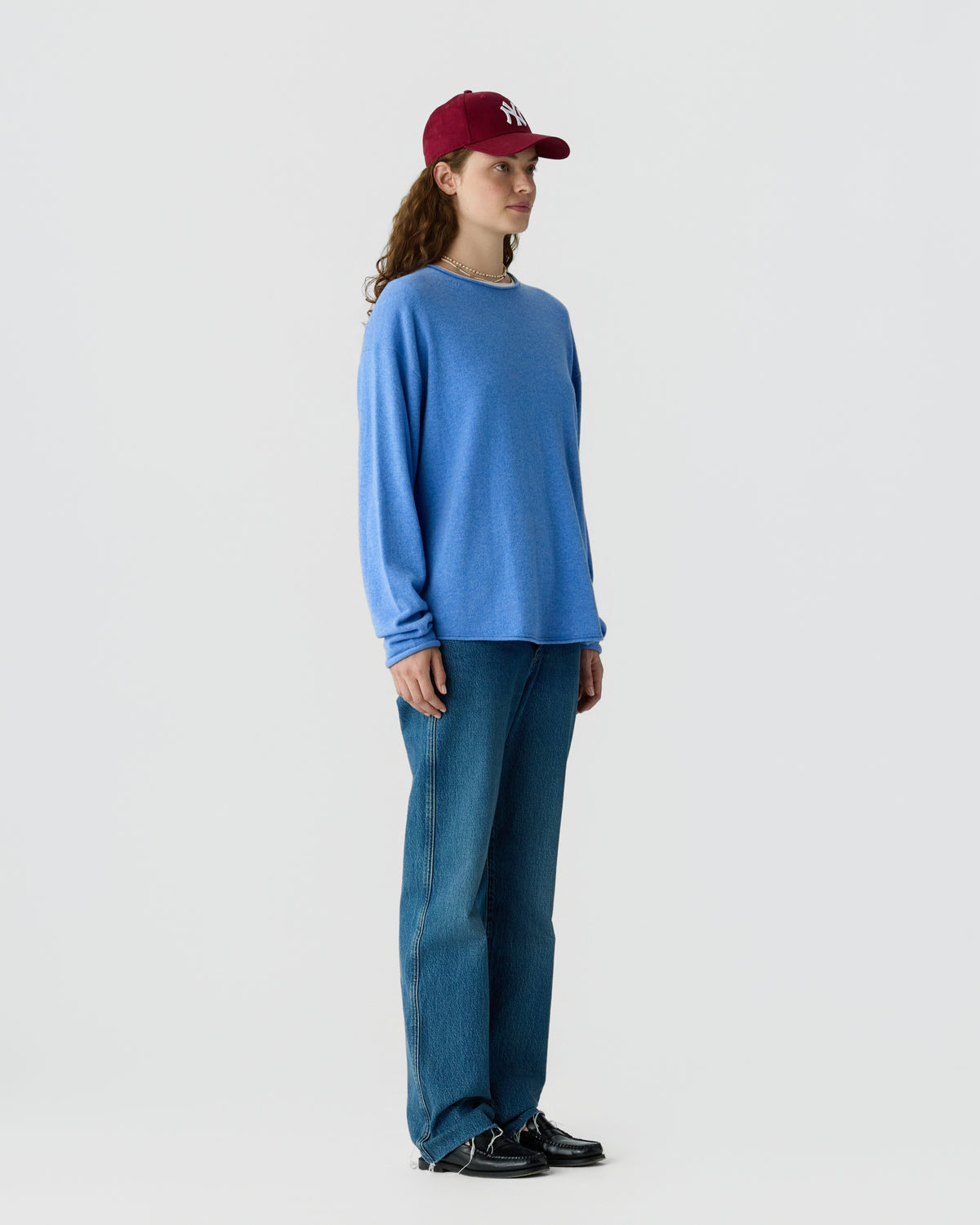 Woman standing in profile wearing a loose blue long-sleeve sweater, blue jeans, black shoes, and a red baseball cap against a plain light gray background.