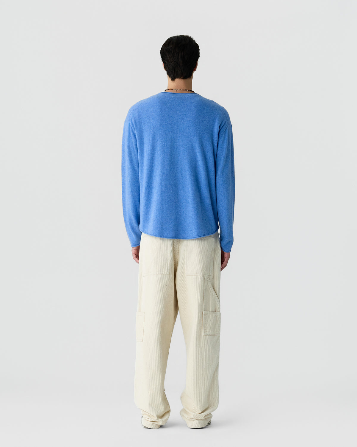 Man standing facing away wearing a light blue long-sleeve sweater and loose cream-colored pants against a plain light gray background.