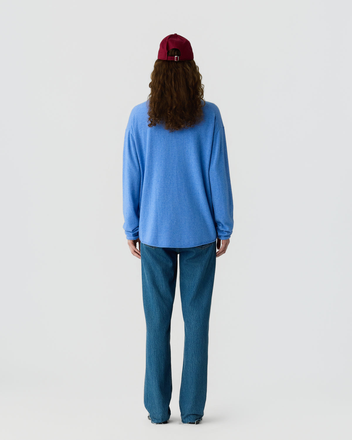 Person with long curly hair wearing a red baseball cap, a loose light blue sweater, and straight blue jeans, standing facing away against a plain white background.