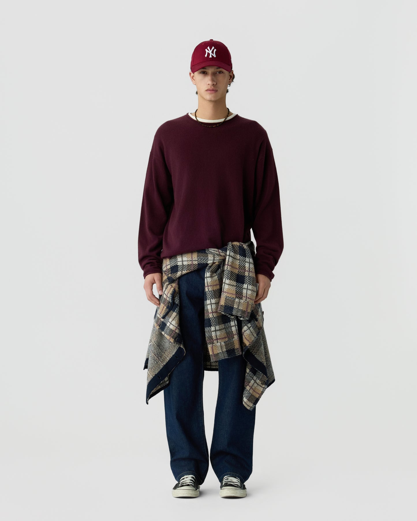 Young man wearing a burgundy long-sleeve sweater, matching burgundy cap, dark blue jeans, and a plaid jacket tied around his waist against a plain light background.